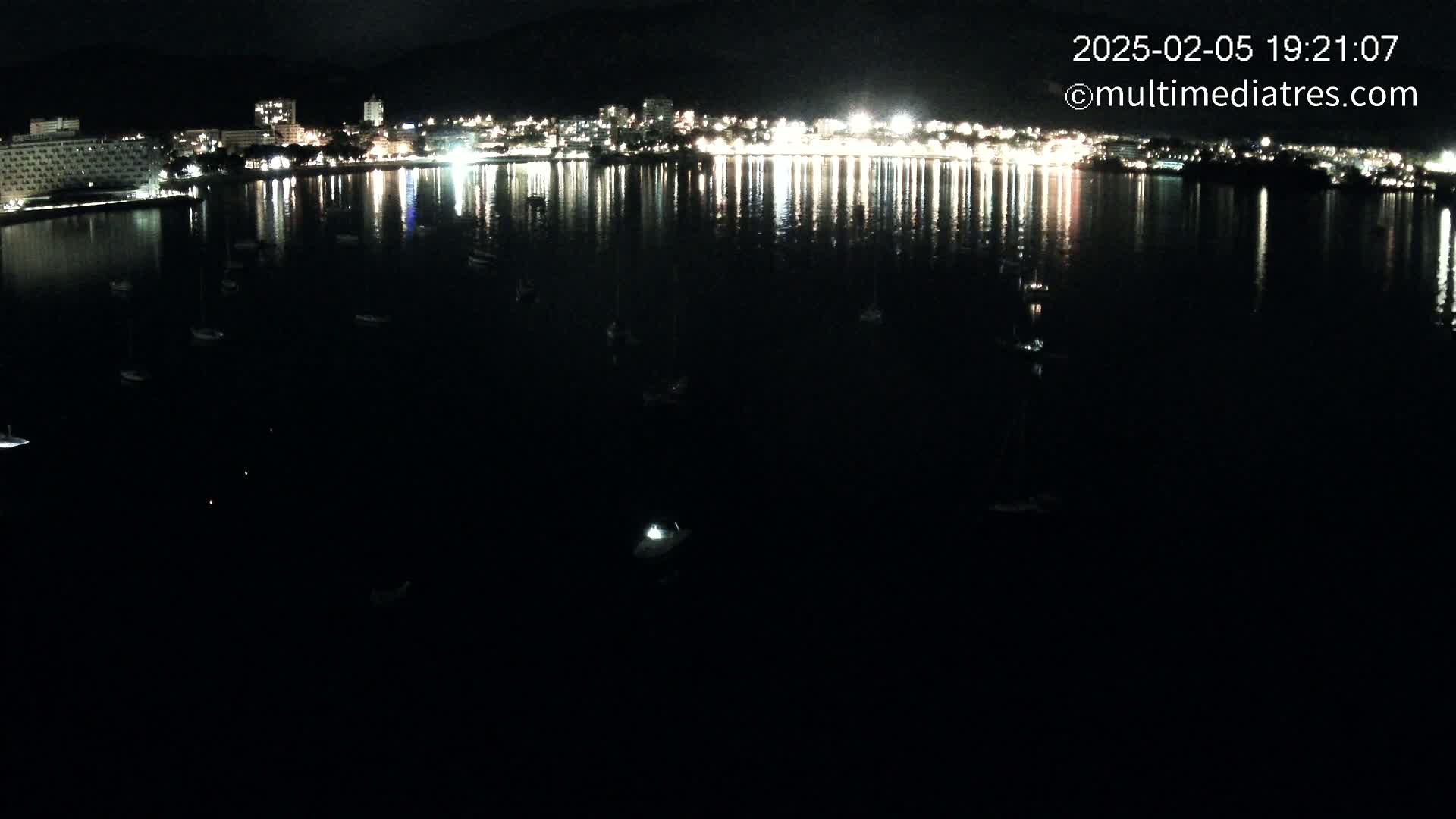 A nighttime view of a calm body of water reflecting the lights of a city on the far shore, with several small boats scattered across the water.