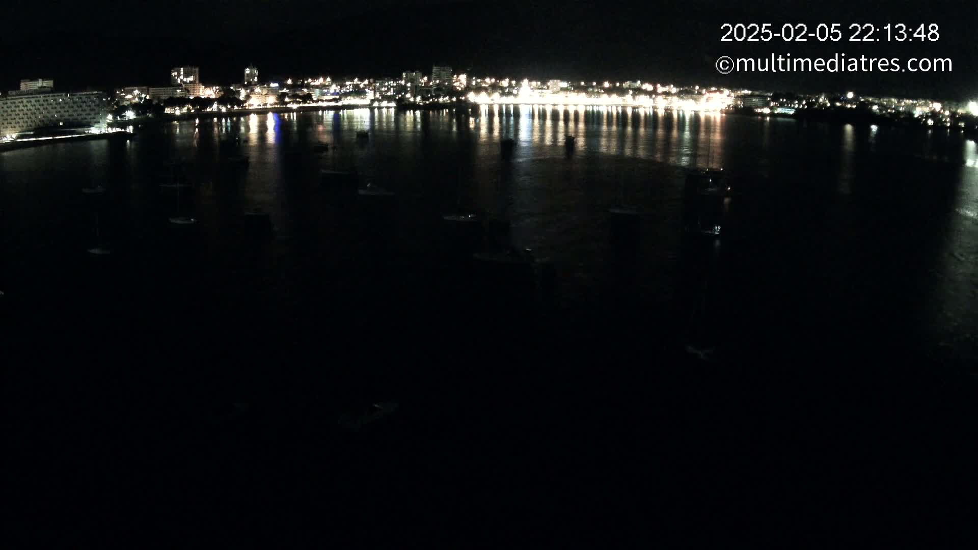 A nighttime view of a calm body of water with numerous boats moored, reflecting the lights of a city along the shoreline.