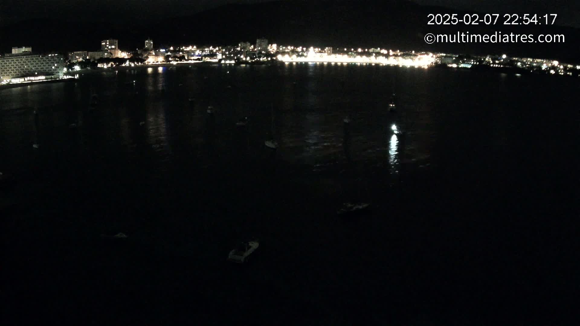 A nighttime, aerial view shows a dark body of water with several small boats, reflecting the lights of a city on the shoreline.