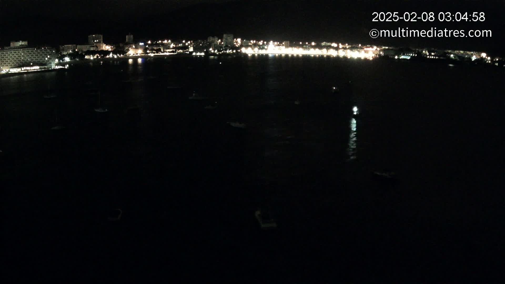 A nighttime, aerial view shows a dark body of water with several small boats, and a brightly lit city skyline in the distance.