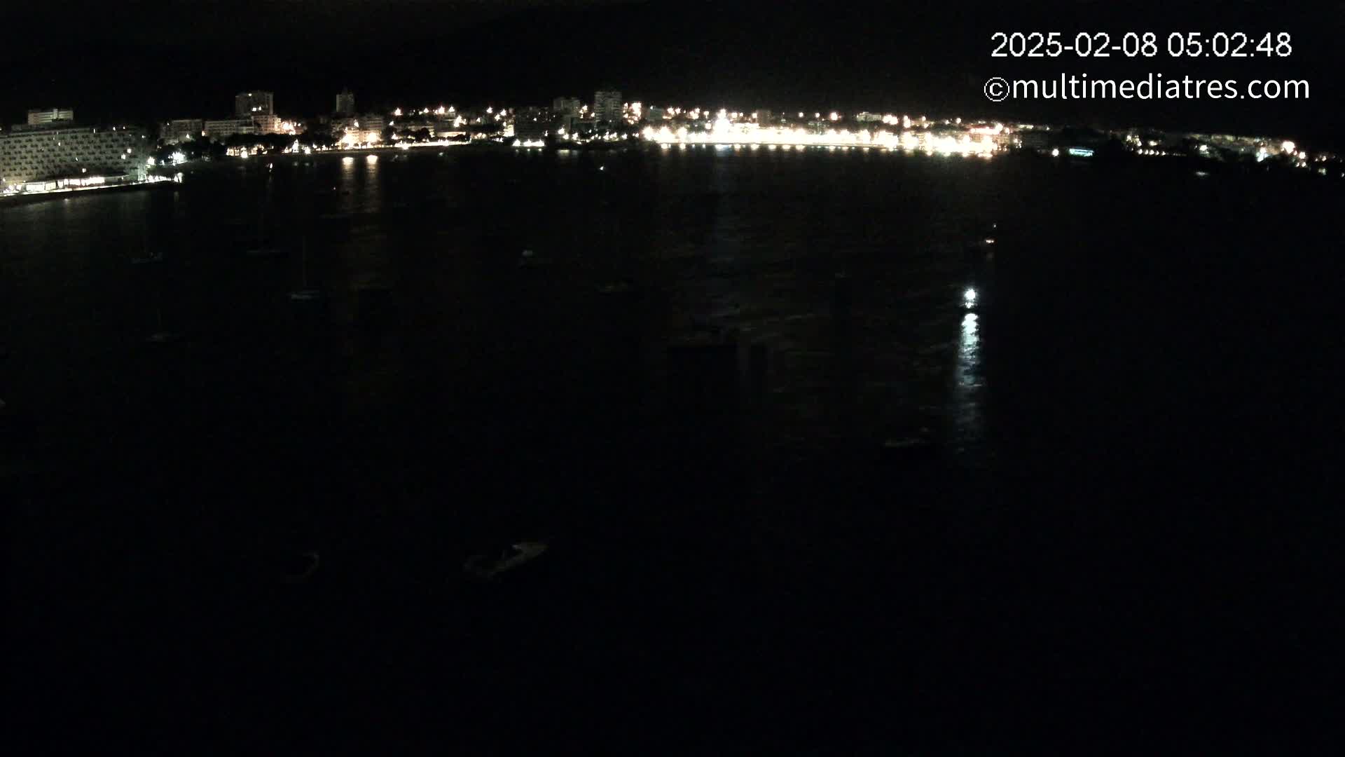 A nighttime, long-range view shows a dark body of water with a few small boats, reflecting the lights of a city's buildings along the shoreline.