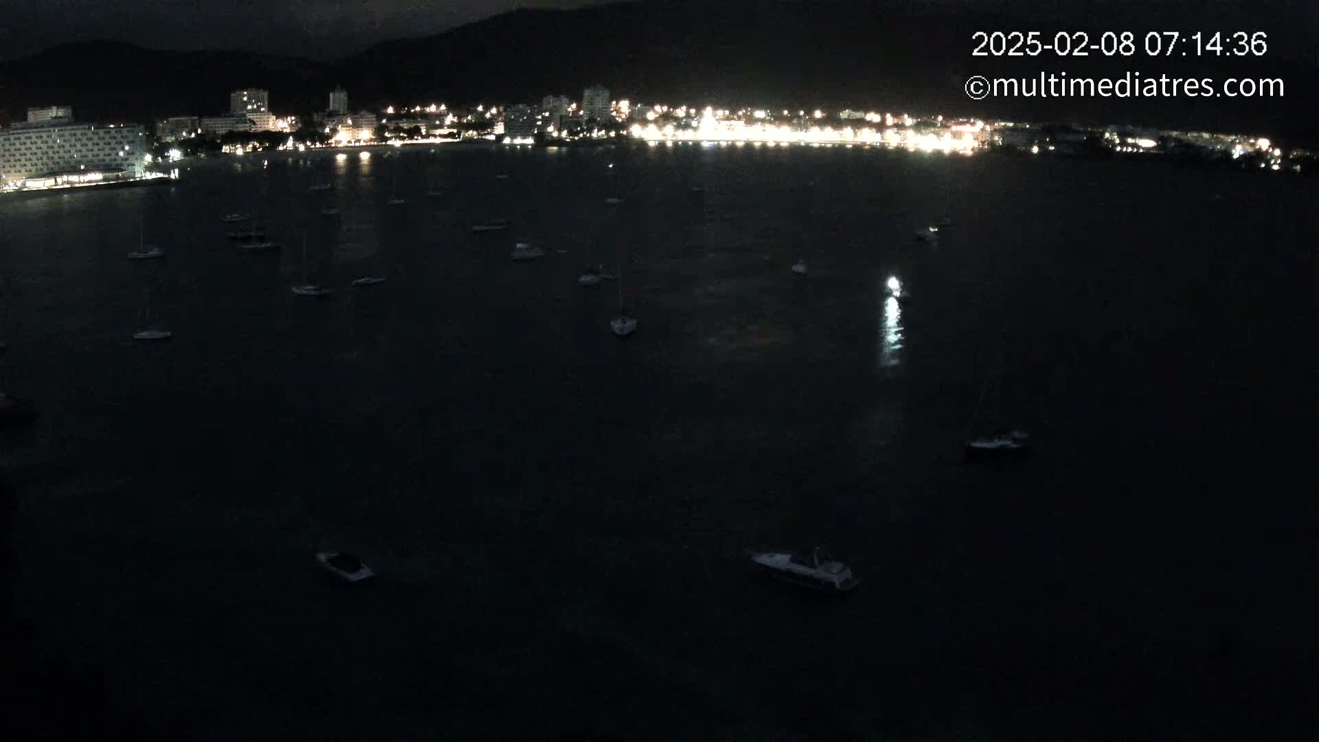 A nighttime view of a dark body of water with numerous boats and a brightly lit coastline in the background.
