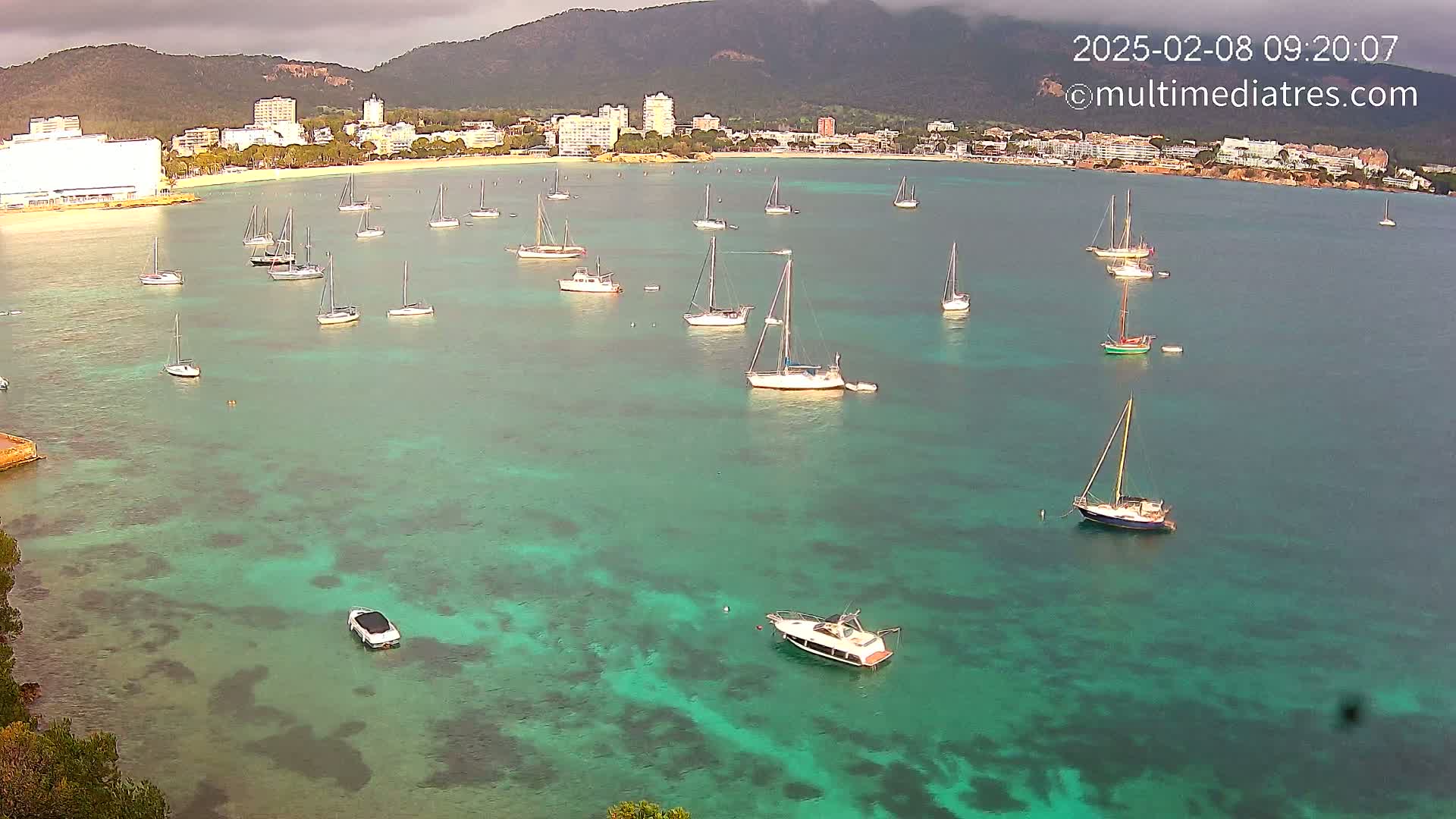 Numerous sailboats are anchored in a calm, clear, teal-colored bay near a coastline with buildings and mountains under an overcast sky.