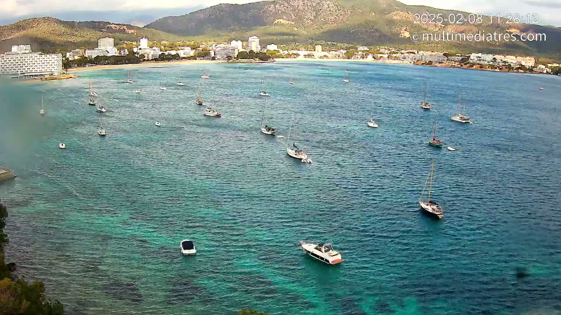 Numerous sailboats and motorboats are anchored in a calm, clear turquoise bay near a shoreline with hotels and mountains in the background under a partly cloudy sky.