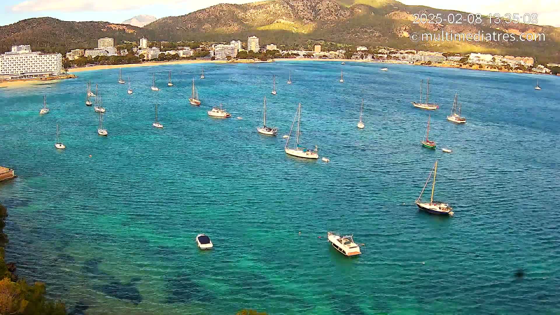 Numerous sailboats are anchored in a calm, clear turquoise bay near a shoreline with hotels and mountains in the background.