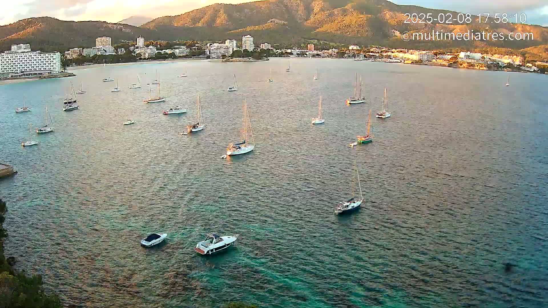 Numerous sailboats are anchored in a calm, teal-colored bay near a coastline with buildings and mountains under a partly cloudy sunset sky.