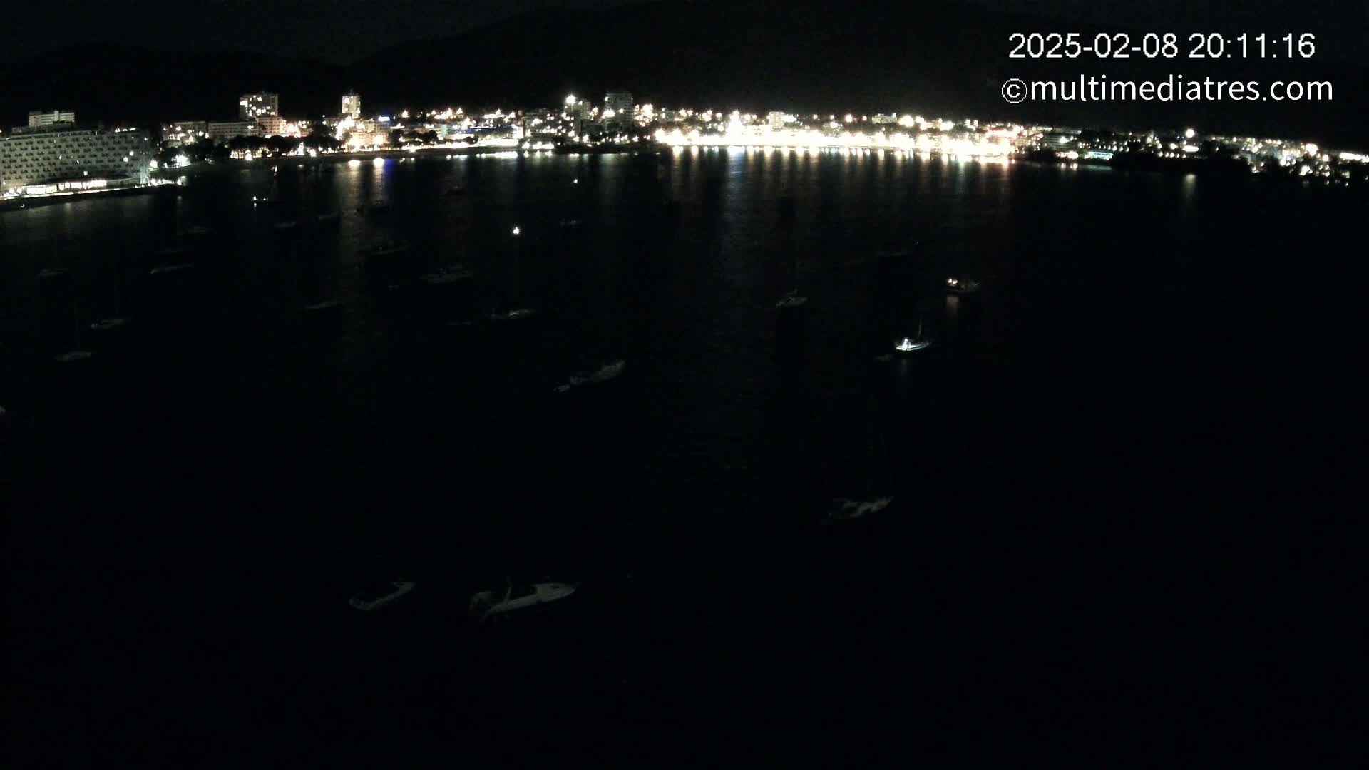 At night, numerous boats are moored in a dark body of water in front of a brightly lit city nestled against a dark mountain.