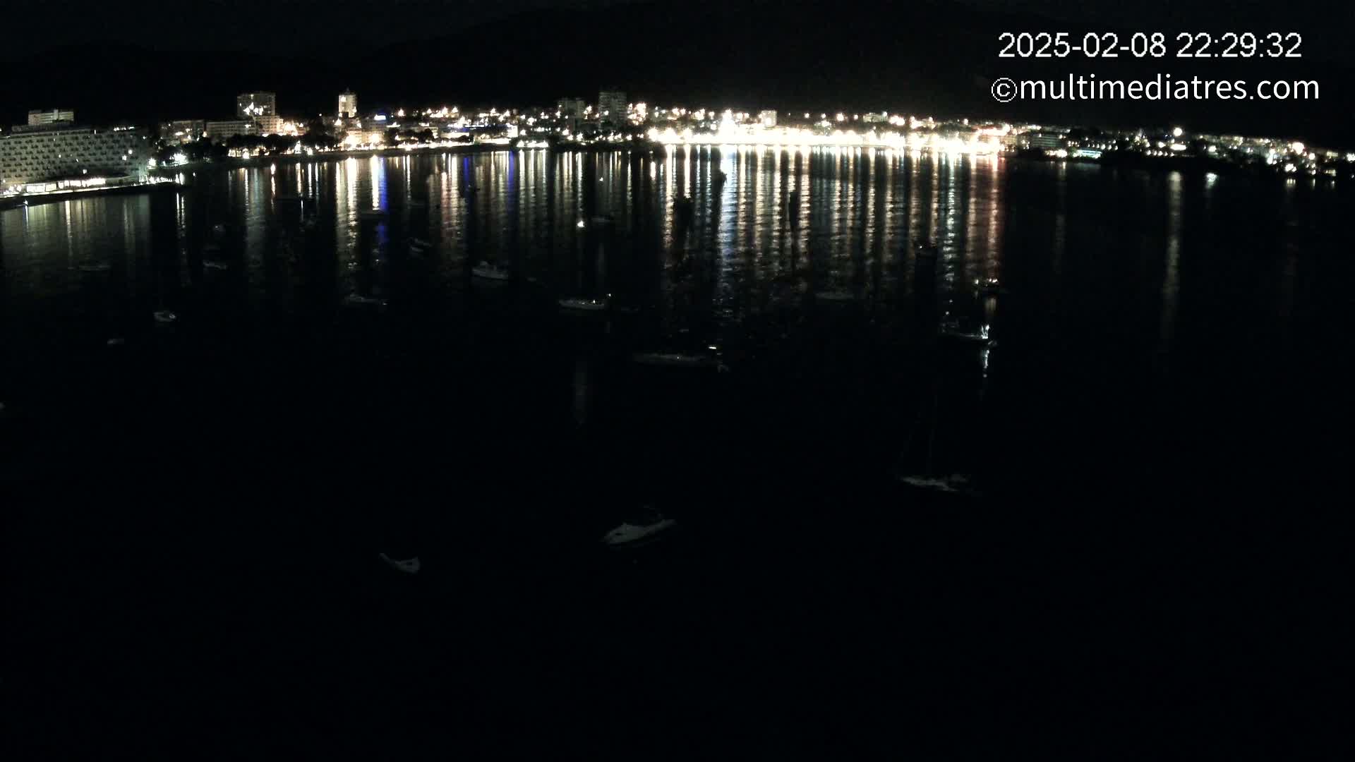 A nighttime view of a city's waterfront, illuminated by lights reflected in calm water, with several small boats scattered across the dark bay.