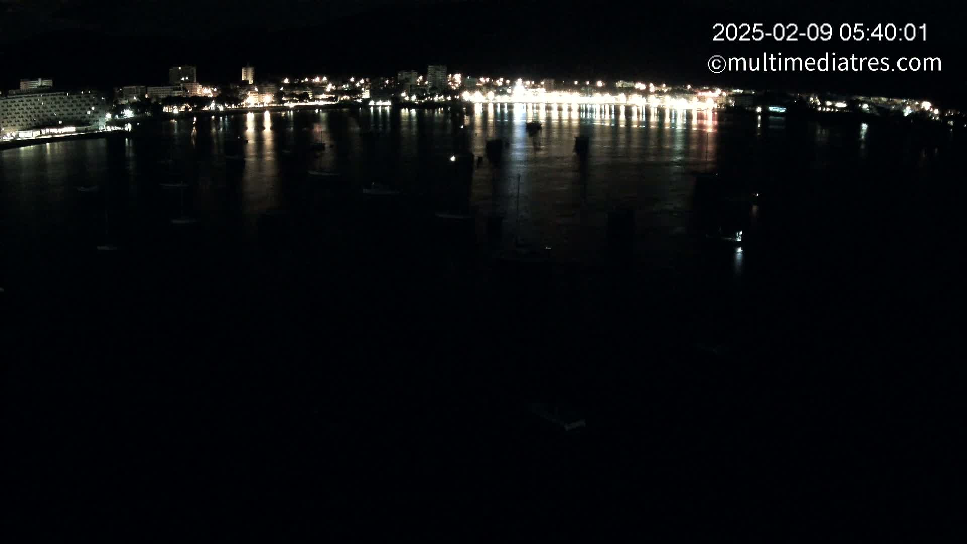 A nighttime view of a city's illuminated waterfront at night, showing numerous boats in a calm body of water.
