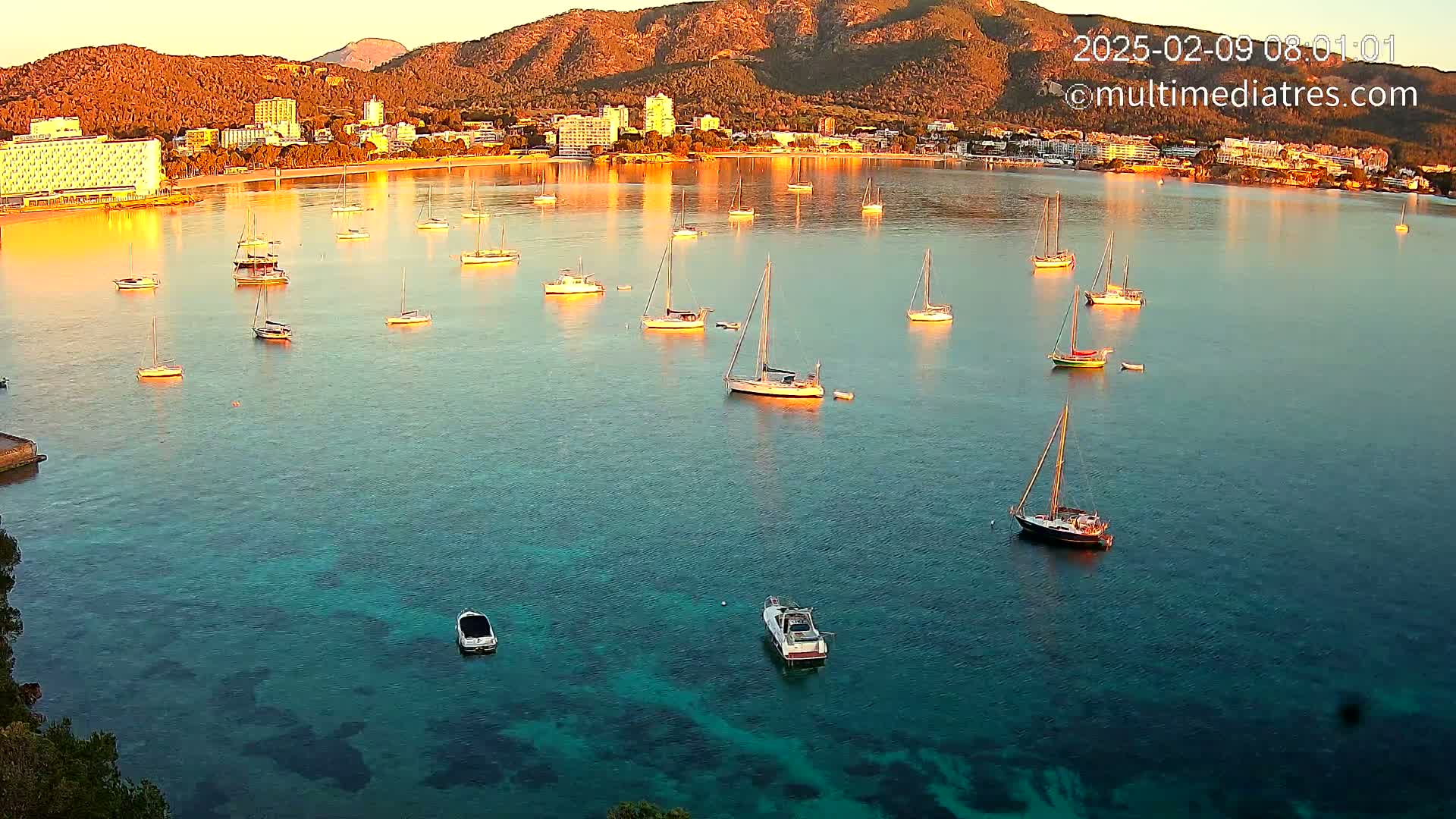 Numerous sailboats are anchored in a calm, clear bay at sunrise, with a town and mountains visible in the background.