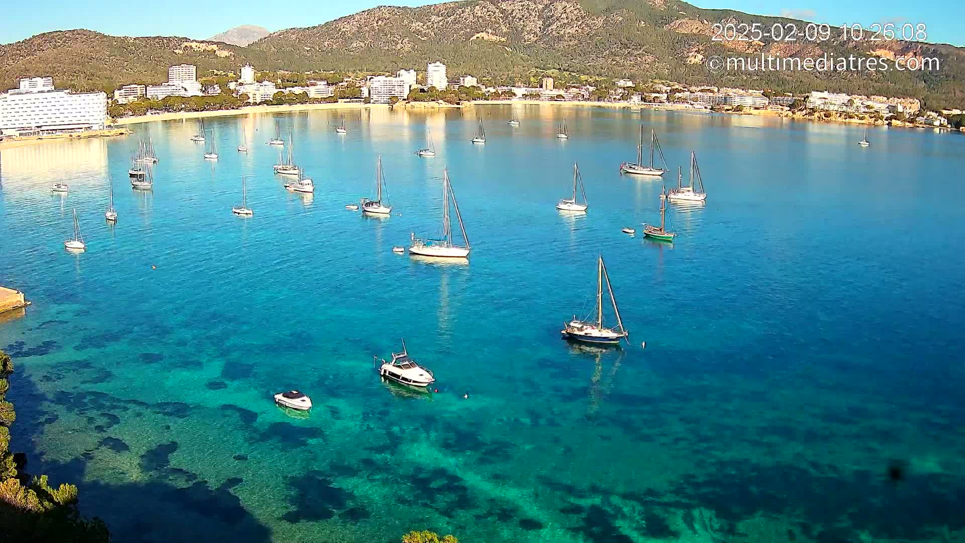 A clear, sunny day shows a calm bay of turquoise water with numerous sailboats and motorboats anchored near a coastline with hotels and mountains in the background.