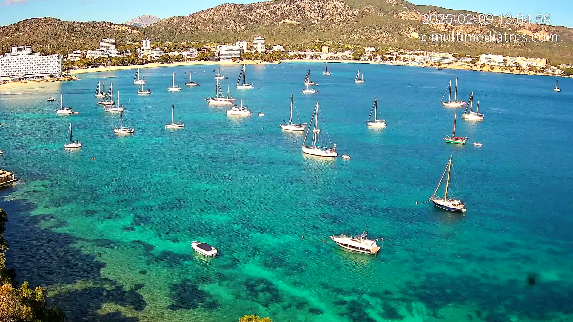Numerous sailboats are anchored in a calm, clear turquoise bay near a beach and hotels situated against a mountainous backdrop under a sunny sky.