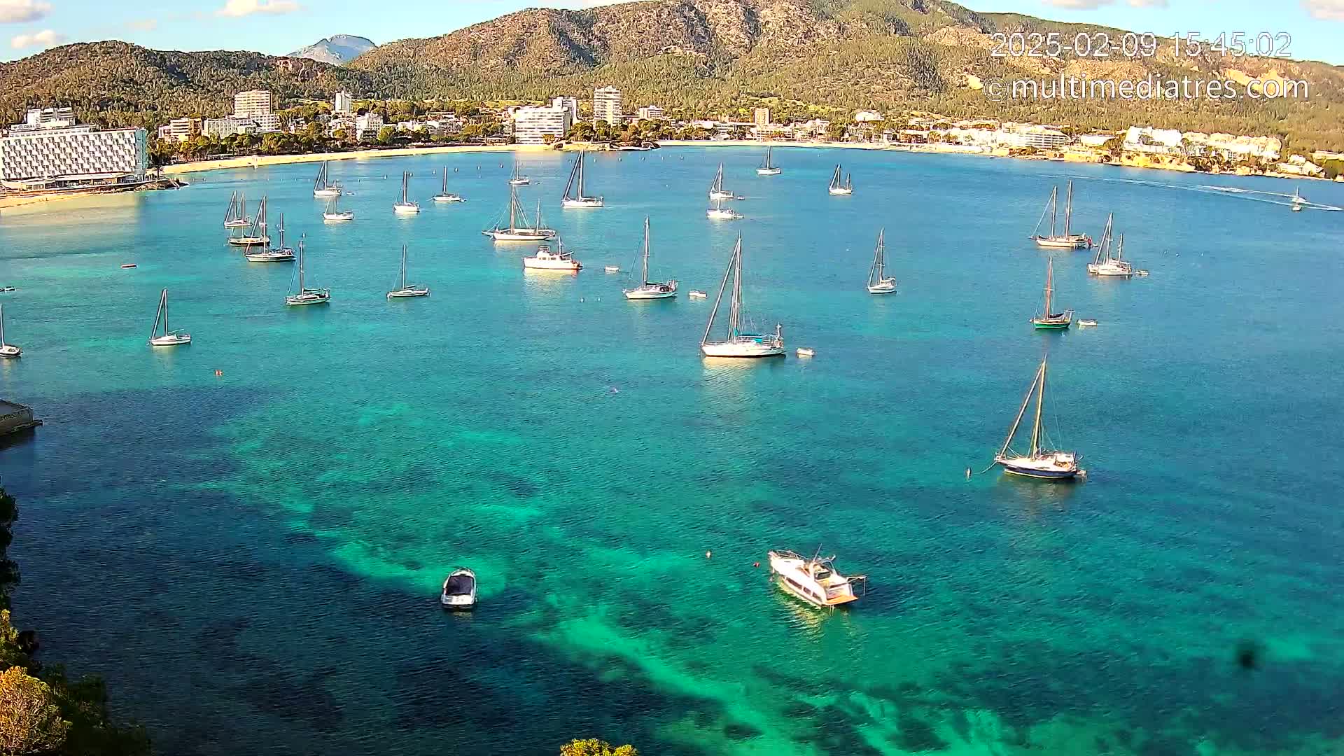 Numerous sailboats are anchored in a calm, clear turquoise bay near a sandy beach and a mountainous coastline under a partly cloudy sky.