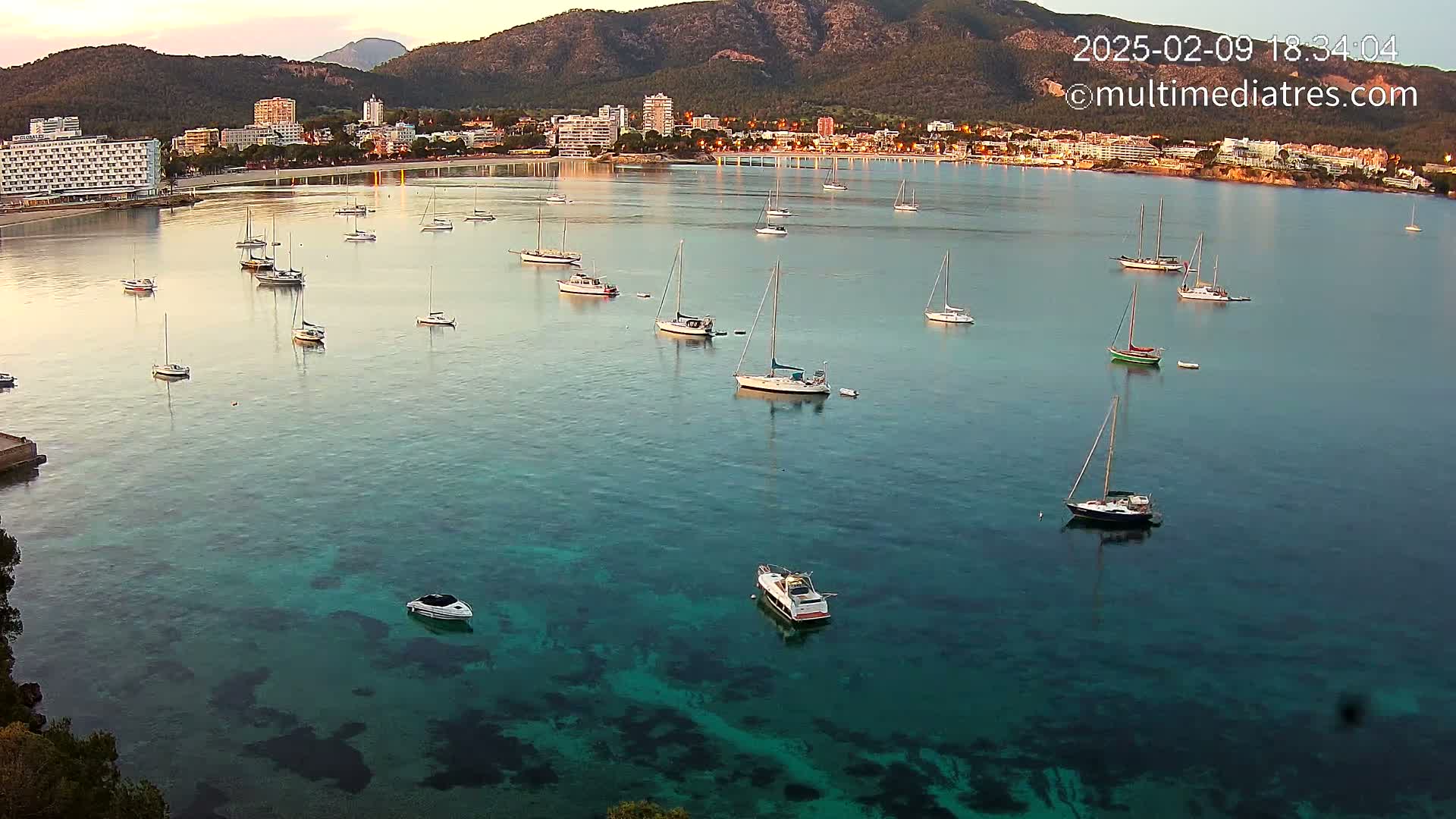 Numerous sailboats and motorboats are anchored in a calm, clear teal bay, with a mountain range and buildings visible in the background under a twilight sky.