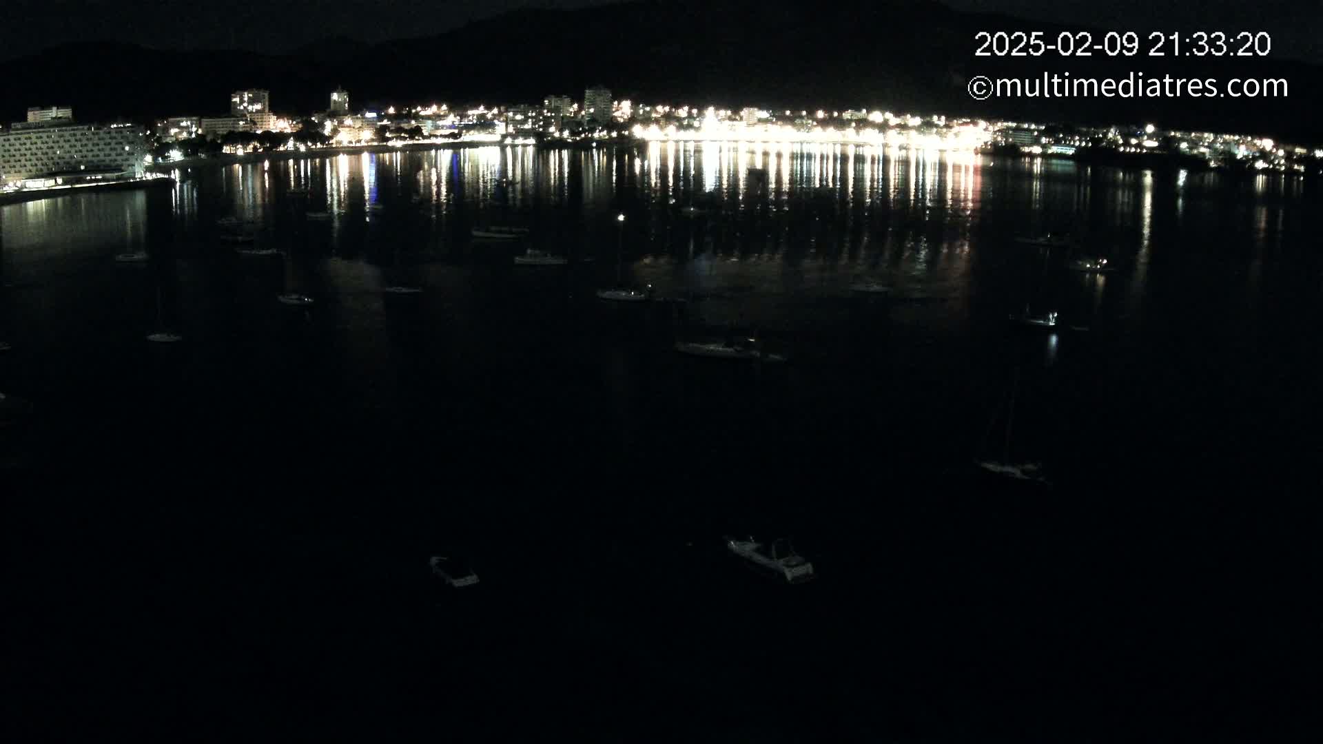 A nighttime view of a calm body of water with numerous small boats, reflecting the lights of a brightly lit city along the shoreline.