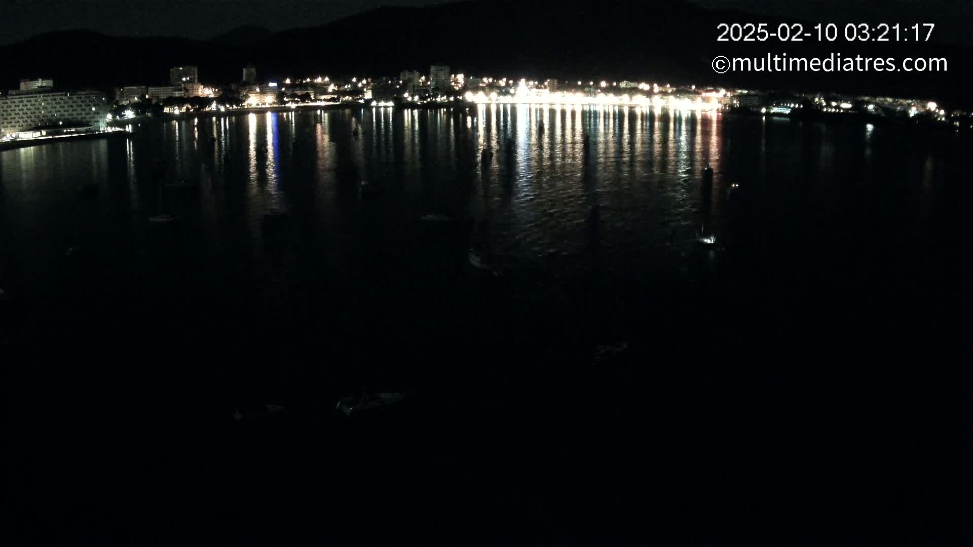 A nighttime view of a calm body of water reflecting the lights of a city on the shore under a dark sky.