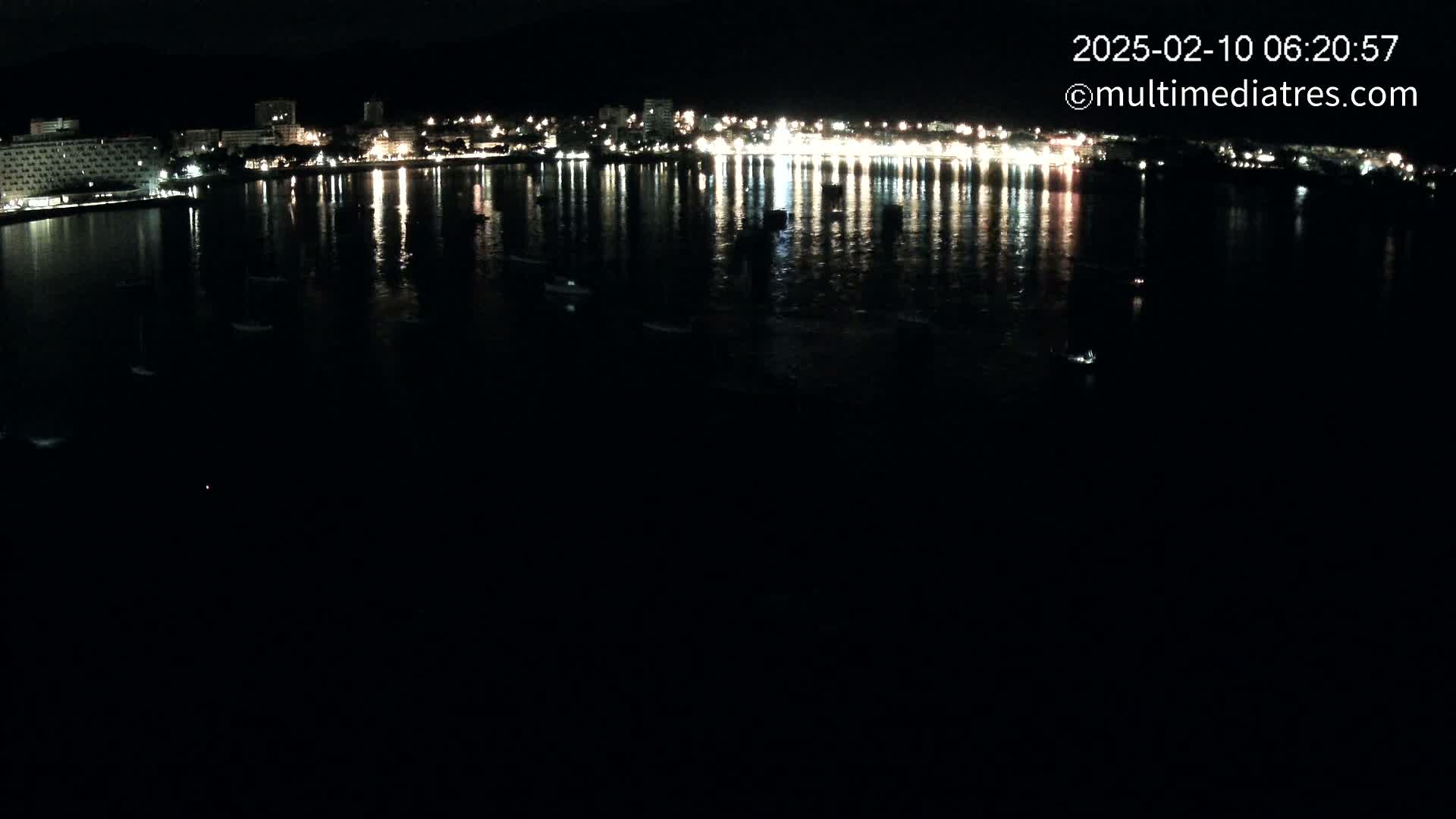 A nighttime view of a city's waterfront, illuminated by lights reflecting on calm water, with several small boats visible.