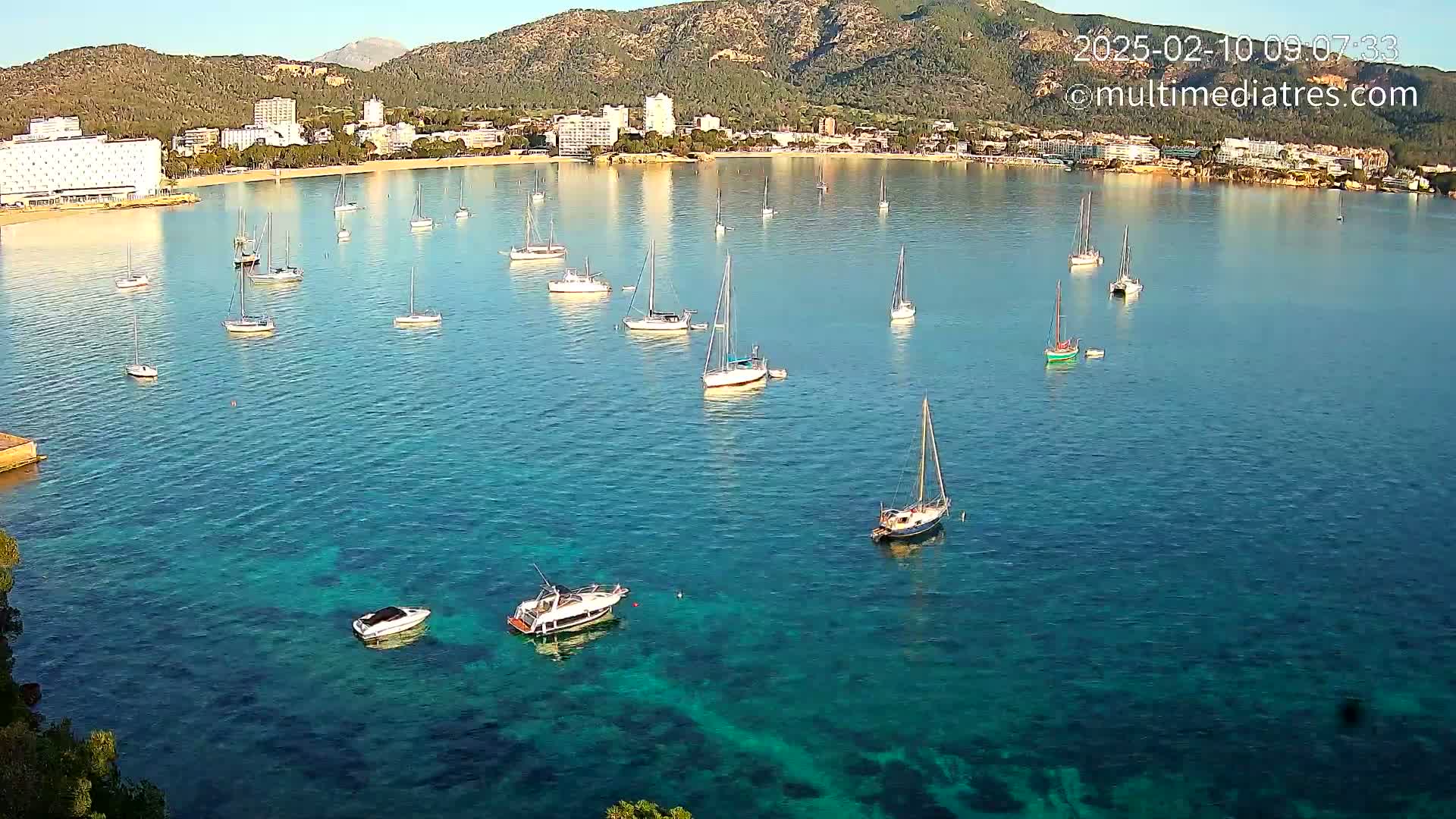Numerous sailboats and motorboats are anchored in a calm, clear turquoise bay near a coastline with hotels and mountains under a sunny sky.
