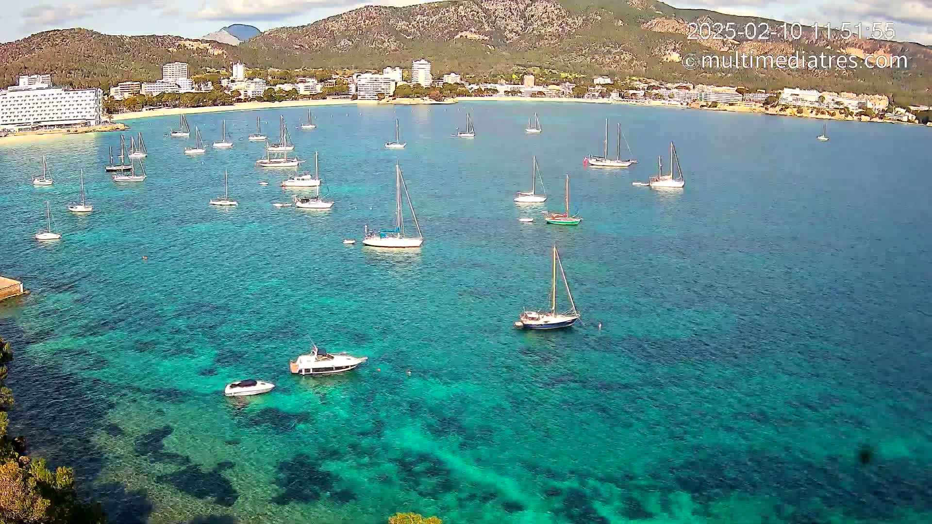 Numerous sailboats and motorboats are anchored in a clear, teal bay near a coastline with hotels and mountains under a mostly sunny sky.