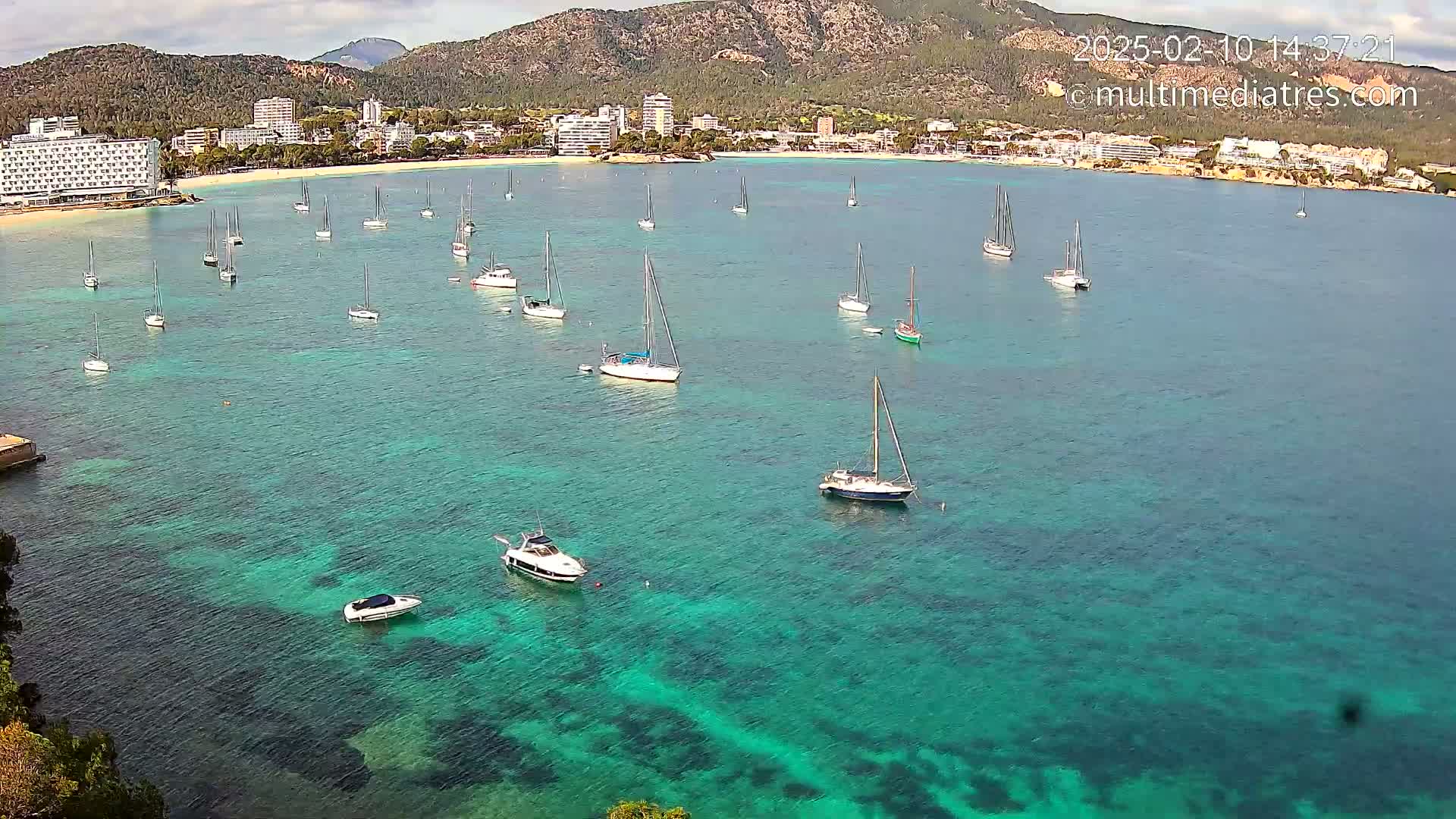 Numerous sailboats and motorboats are anchored in a calm, clear, teal-colored bay near a beach and mountainous coastline.