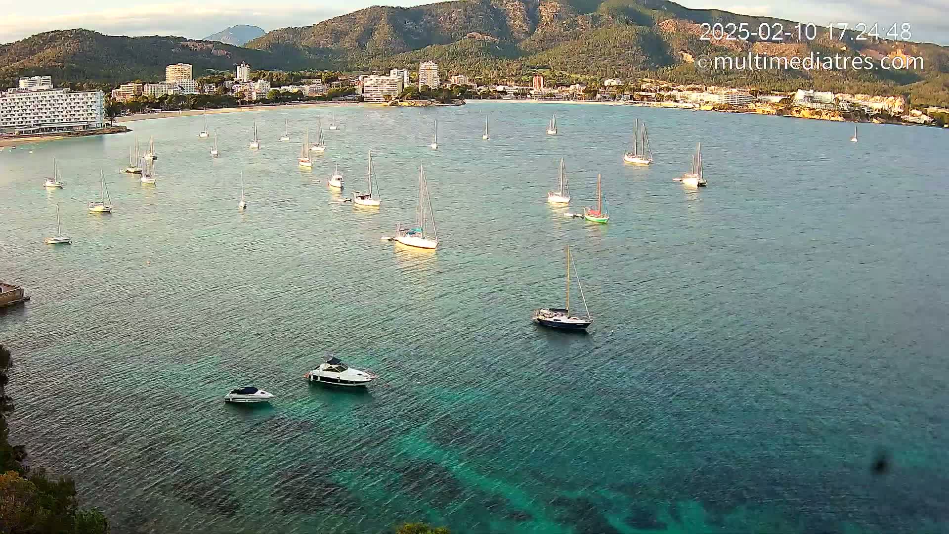 Numerous sailboats and motorboats are anchored in a calm, clear turquoise bay, with a mountainous coastline and buildings in the background.