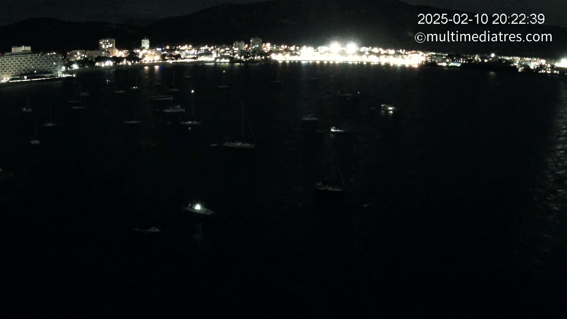 A nighttime, dark-water harbor scene shows many small boats moored near a brightly lit coastal town nestled against dark mountains.