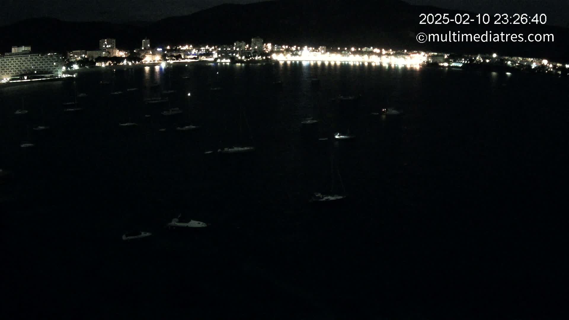 At night, numerous boats are moored in a dark body of water in front of a brightly lit city situated against a dark mountain.