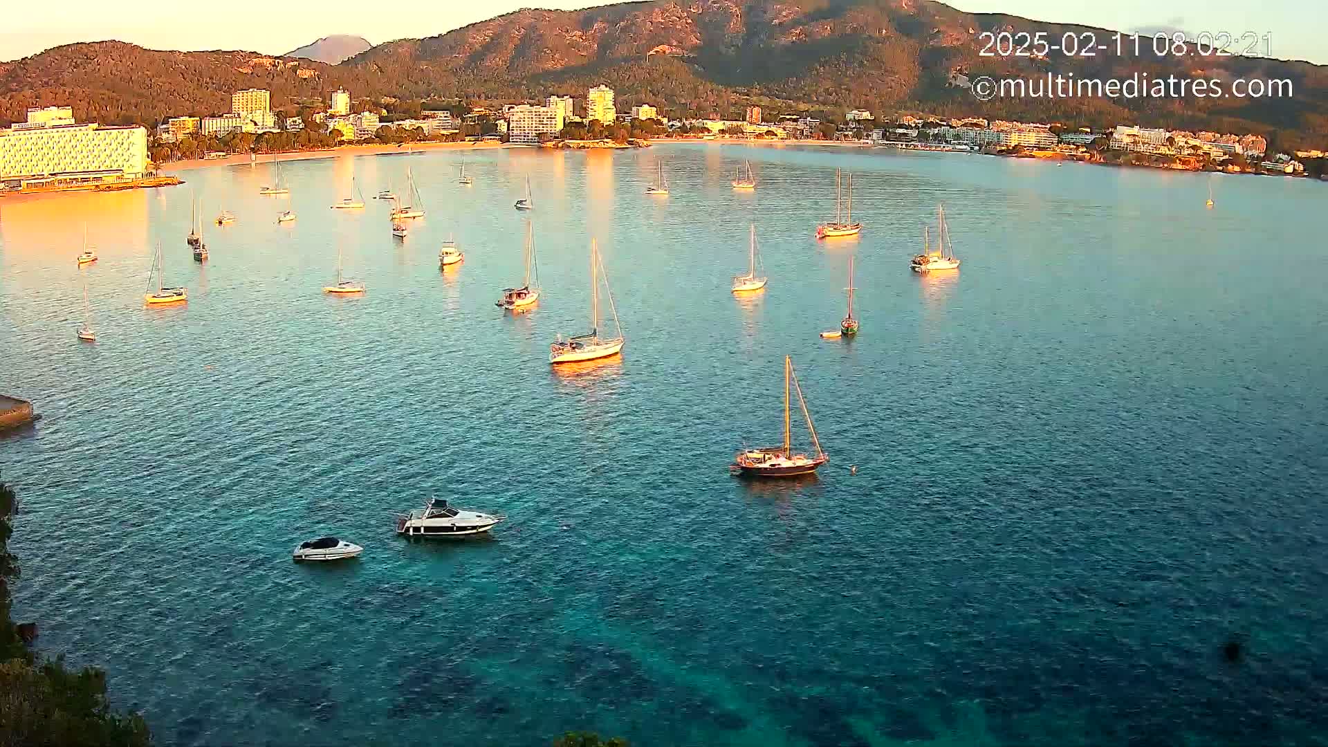 Numerous sailboats and motorboats are anchored in a calm, clear turquoise bay near a shoreline with hotels and mountains in the background under a sunrise or sunset sky.