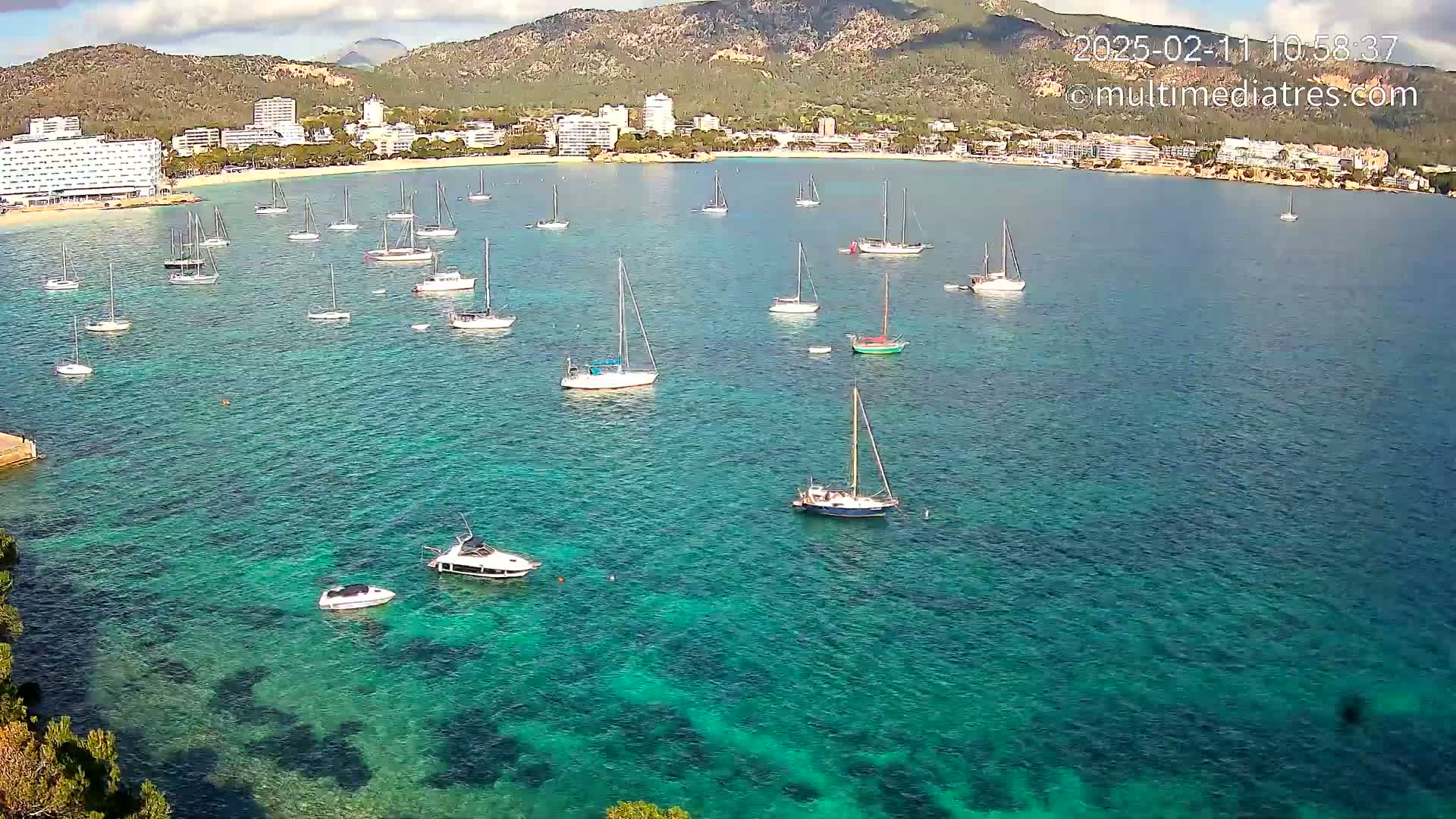 A calm, clear turquoise bay with numerous sailboats and motorboats anchored near a shoreline of hotels and buildings nestled against a mountainous backdrop under a mostly clear sky.