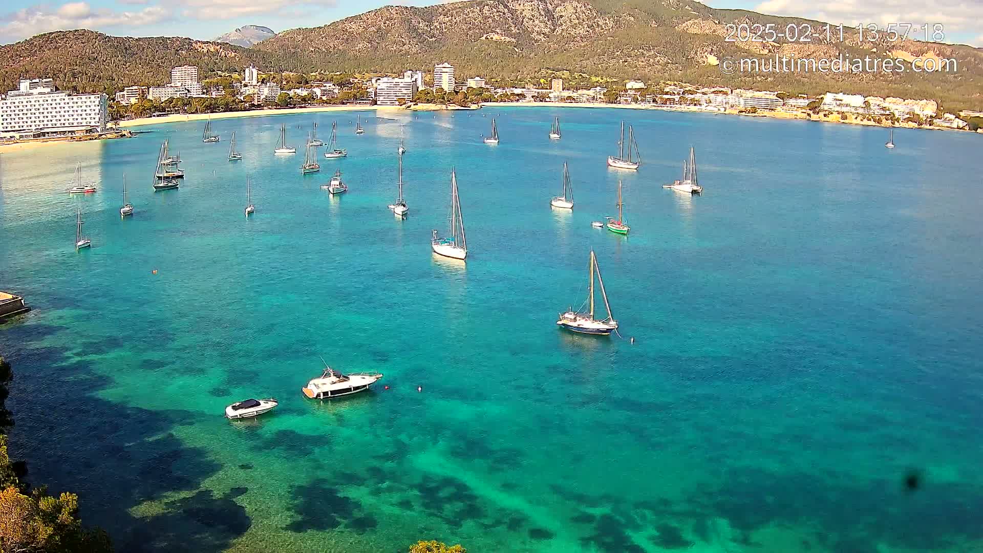 A calm, sunny day shows numerous sailboats and motorboats anchored in a clear, turquoise bay, with a mountainous coastline and hotels in the background.
