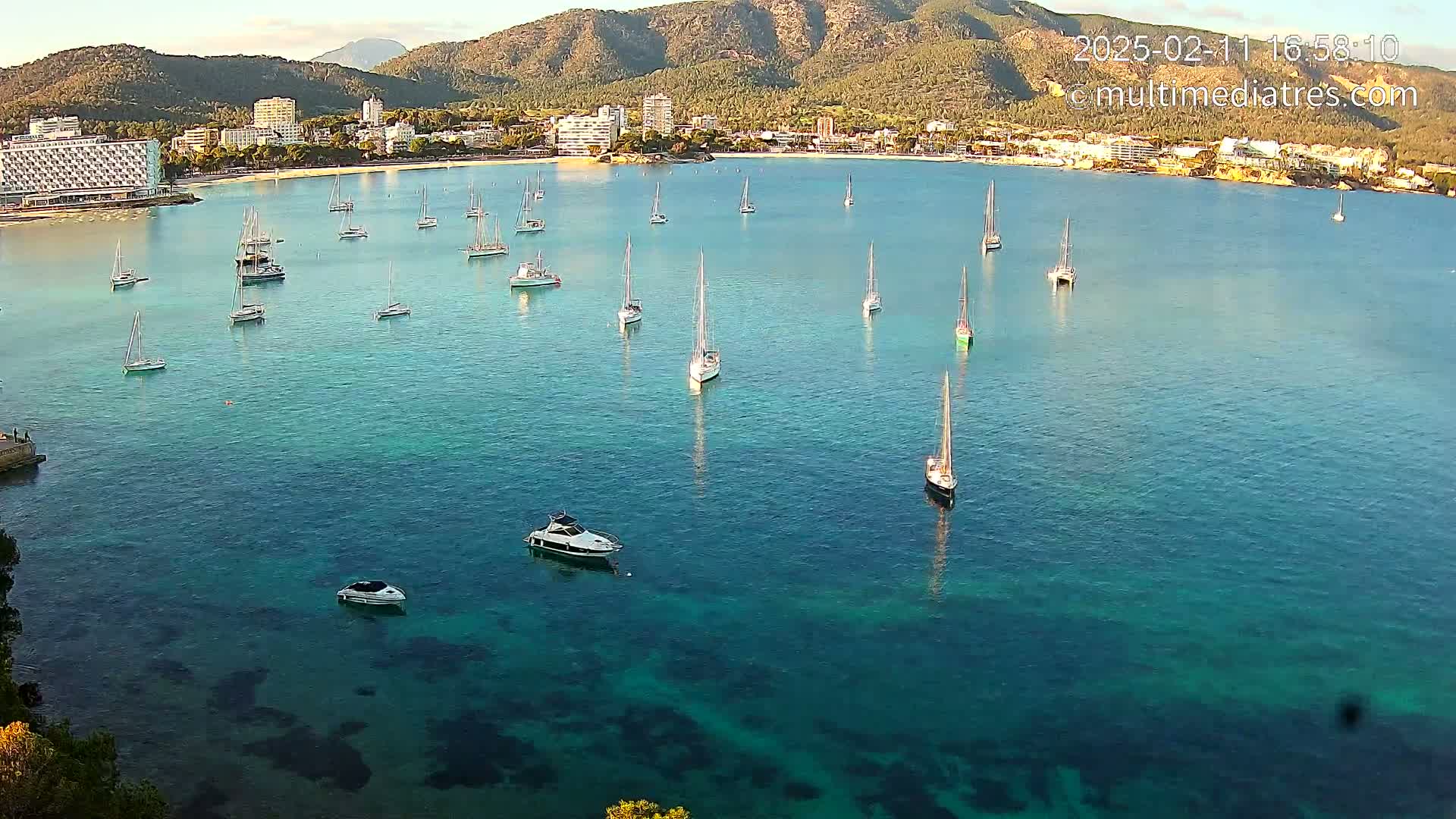 Numerous sailboats and motorboats are anchored in a calm, clear turquoise bay near a shoreline with hotels and mountains in the background under a sunny sky.