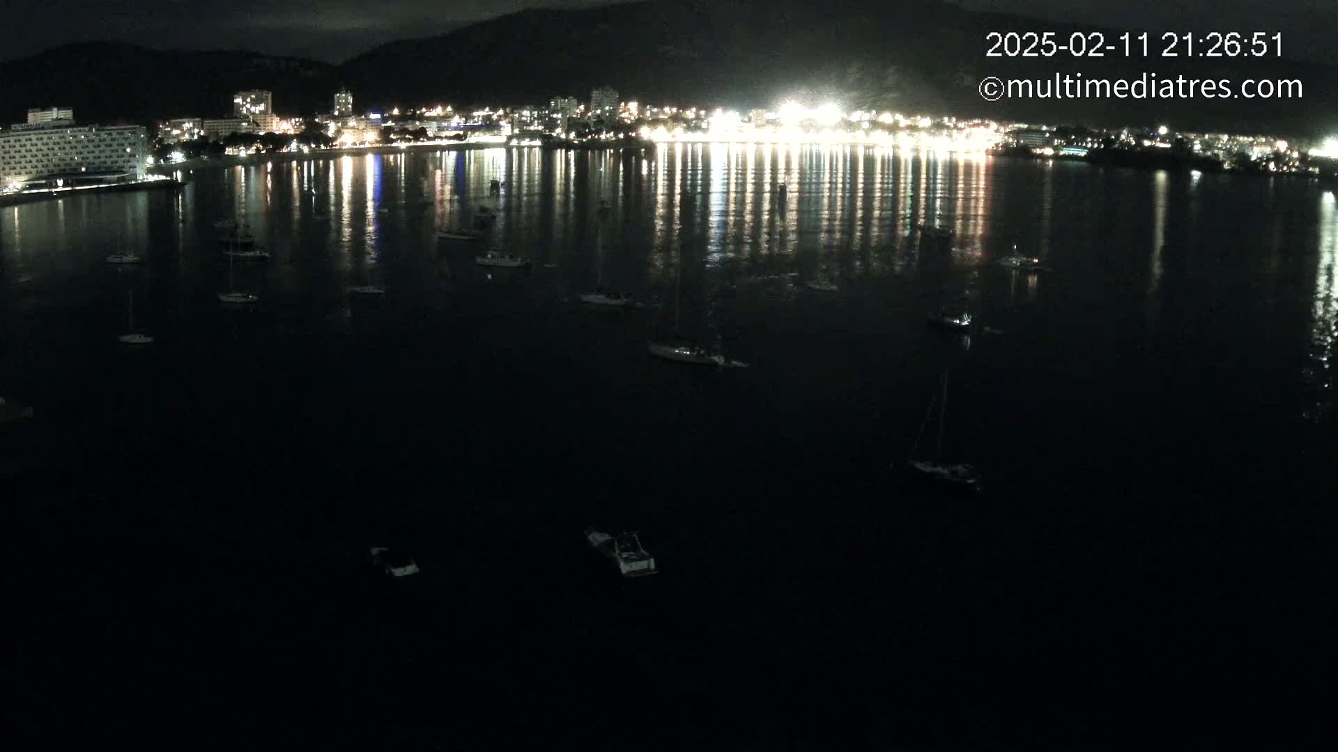 A nighttime view of a calm bay with several small boats moored, reflecting the lights of a city on the far shore.
