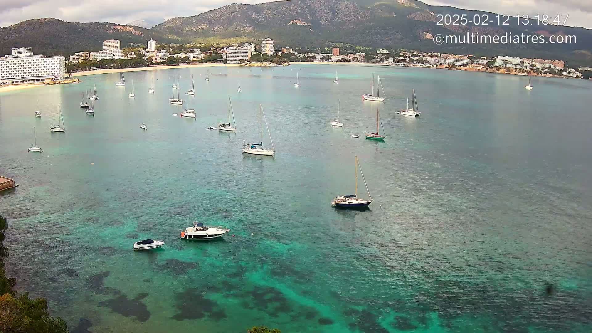 Numerous sailboats and motorboats are anchored in a calm, teal bay near a shoreline with hotels and mountains under a cloudy sky.