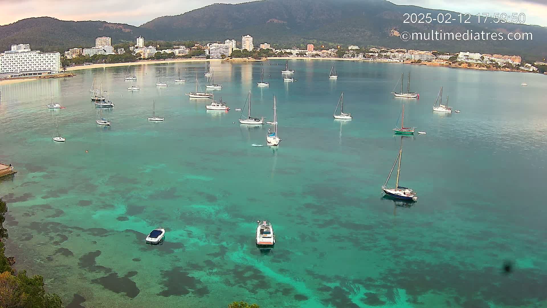 Numerous sailboats are anchored in a calm, clear, teal bay near a coastline with hotels and mountains in the background.