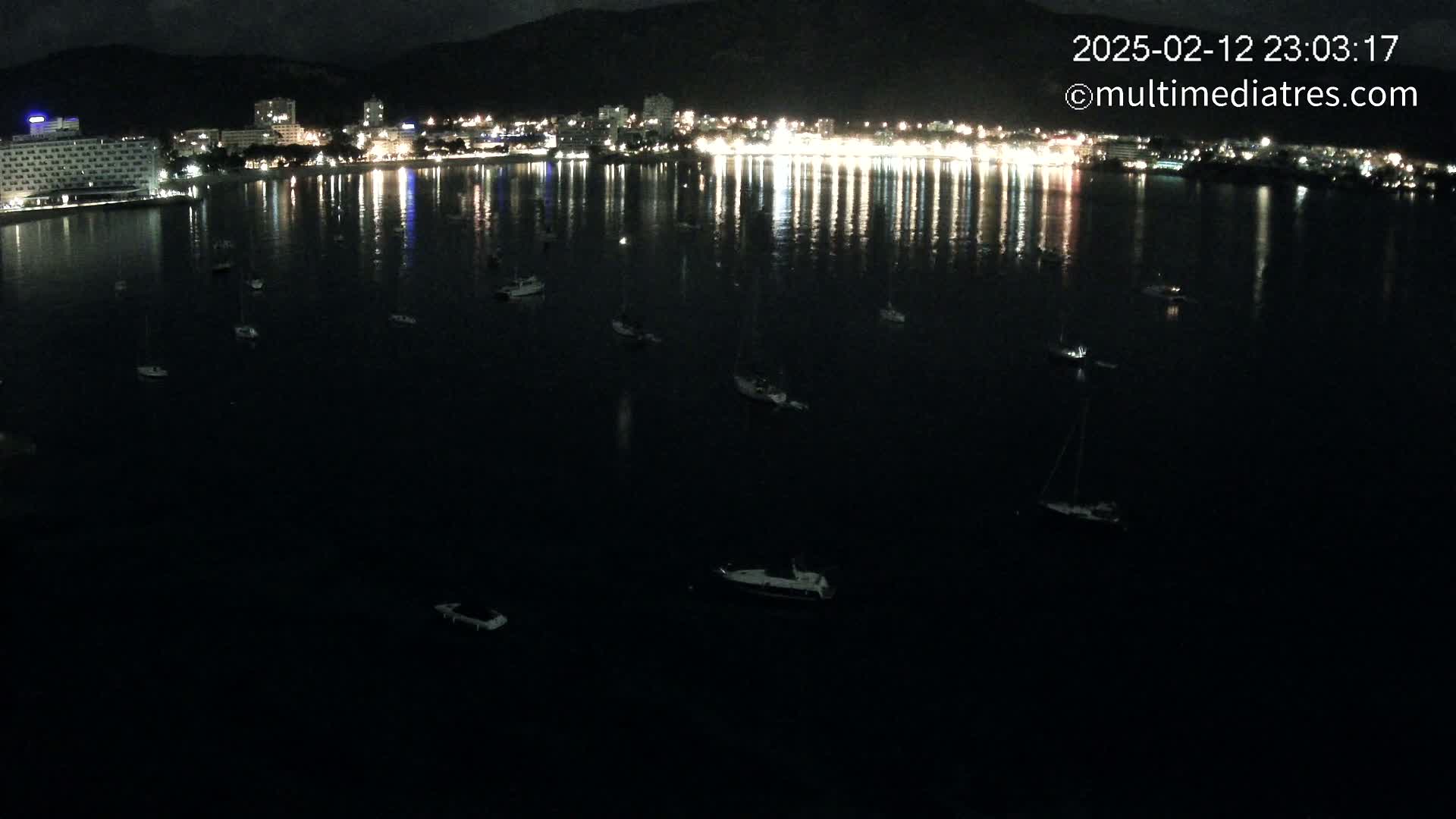 A nighttime view of a calm bay with numerous small boats moored, reflecting the lights of a brightly lit city on the shore.