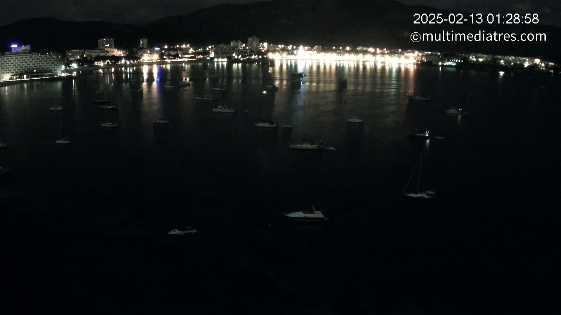 At night, numerous boats are moored in a dark bay in front of a brightly lit coastal town nestled against dark hills.