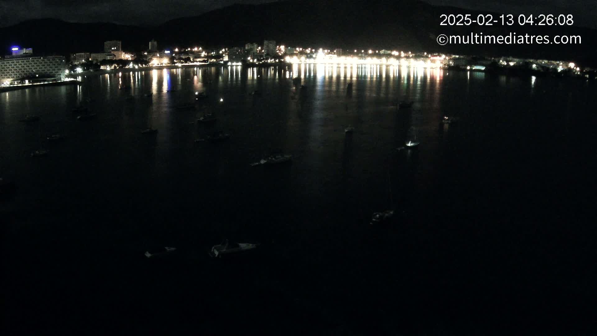A nighttime view of a calm body of water with numerous small boats scattered across its surface, reflecting the lights of a brightly lit city on the far shore, backed by dark mountains.