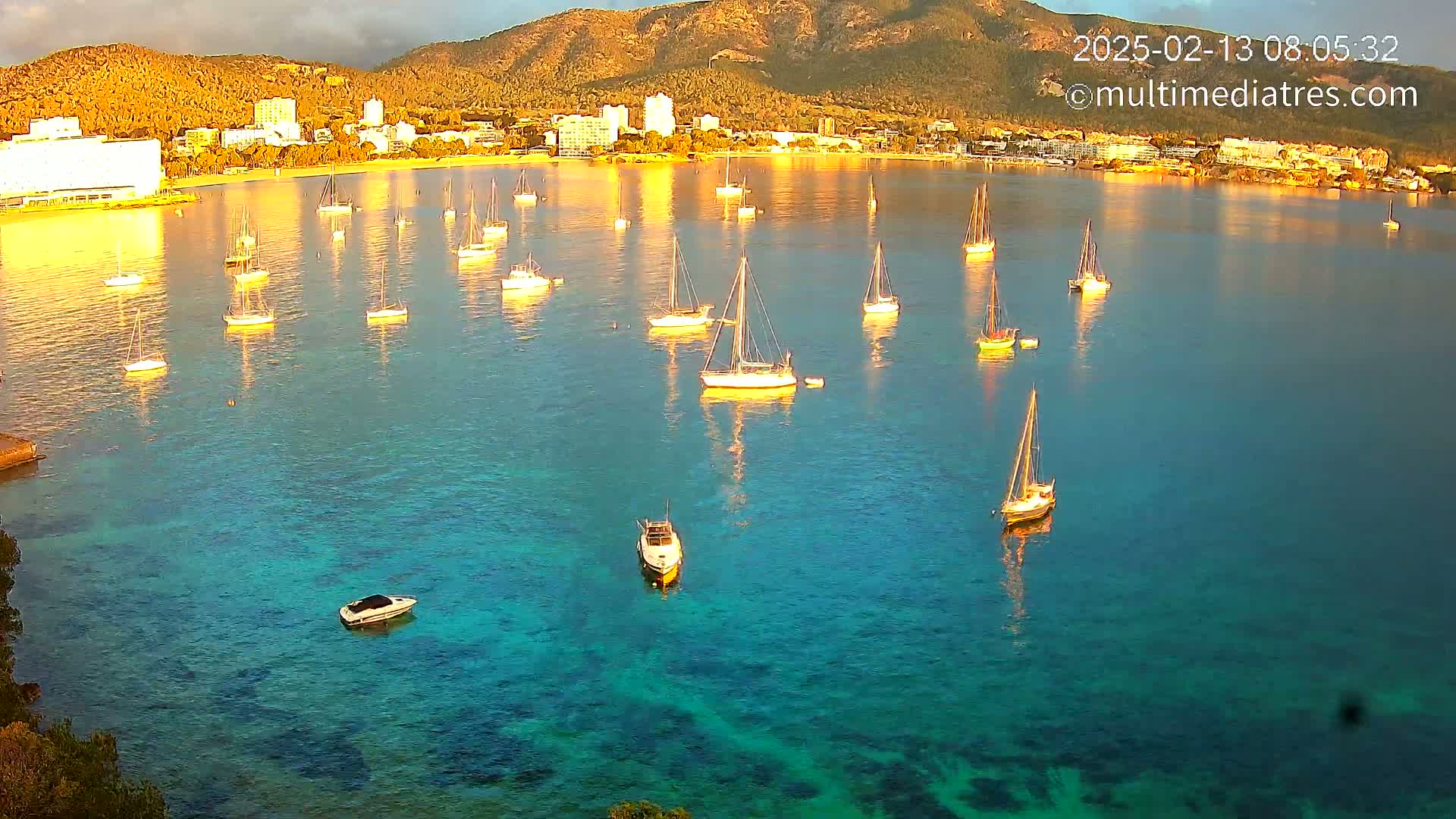 Numerous sailboats and motorboats are anchored in a calm, clear turquoise bay, with a mountain and coastal buildings visible in the background under a partly cloudy sky.