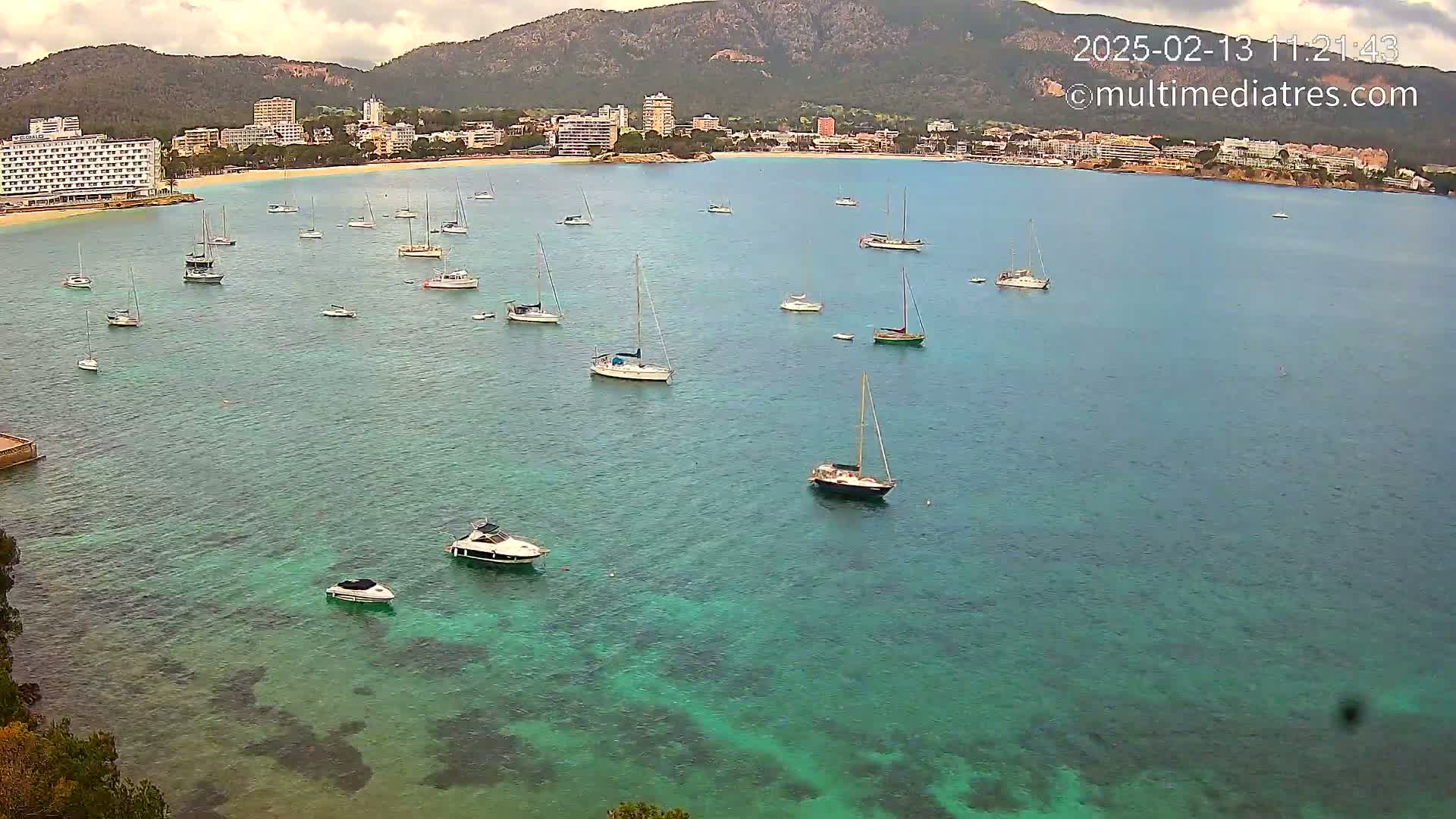 Numerous sailboats and motorboats are anchored in a calm, clear turquoise bay, with a mountainous coastline and hotels visible in the background under partly cloudy skies.