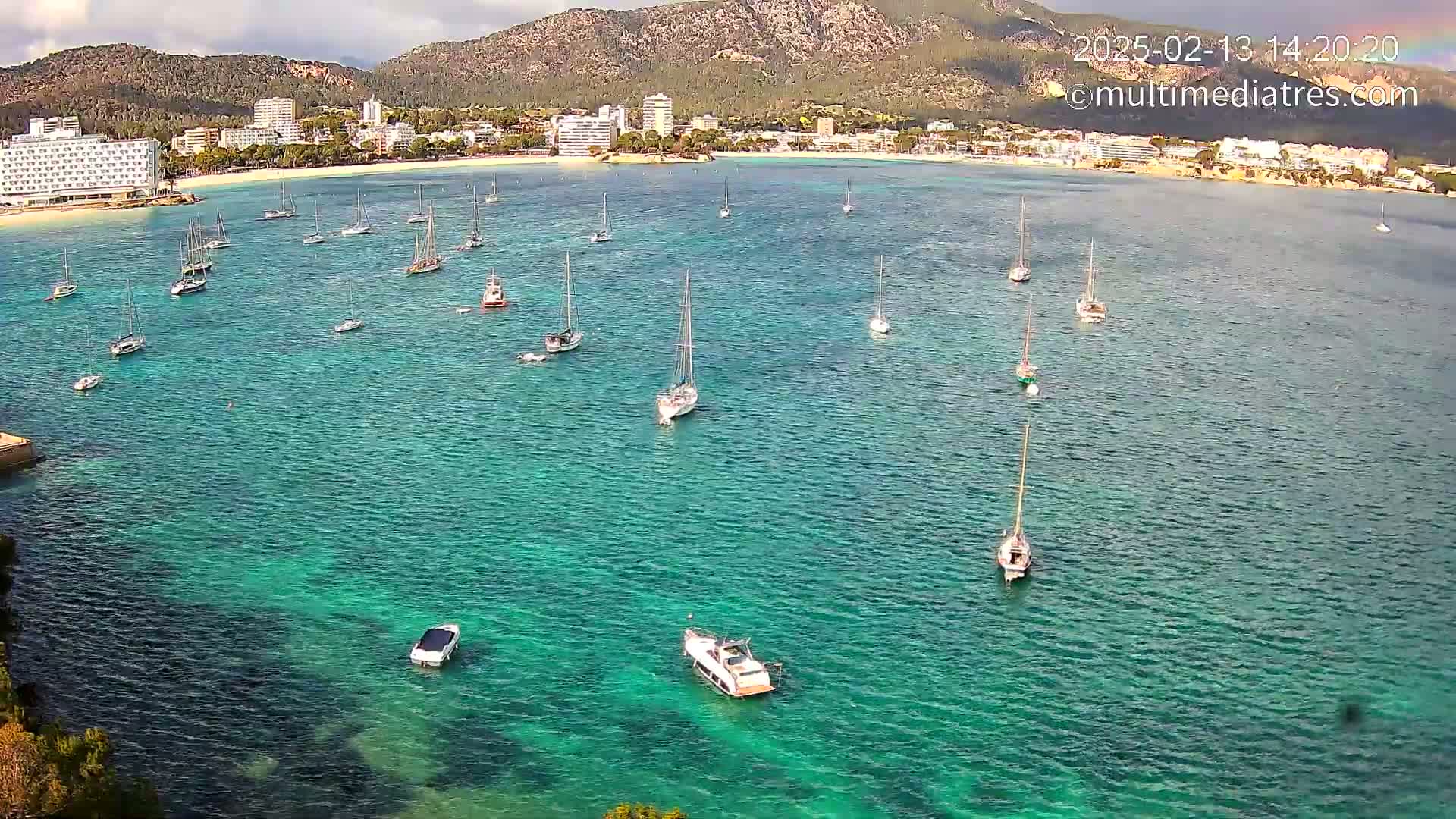 A calm, turquoise bay with numerous sailboats and motorboats anchored near a beach and town backed by mountains under a partly cloudy sky.