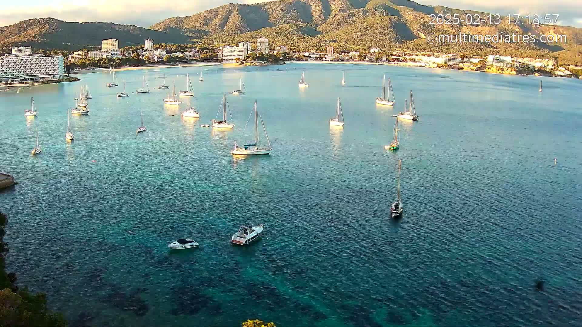 Numerous sailboats are anchored in a calm, clear bay near a mountainous coastline under a mostly clear sky.