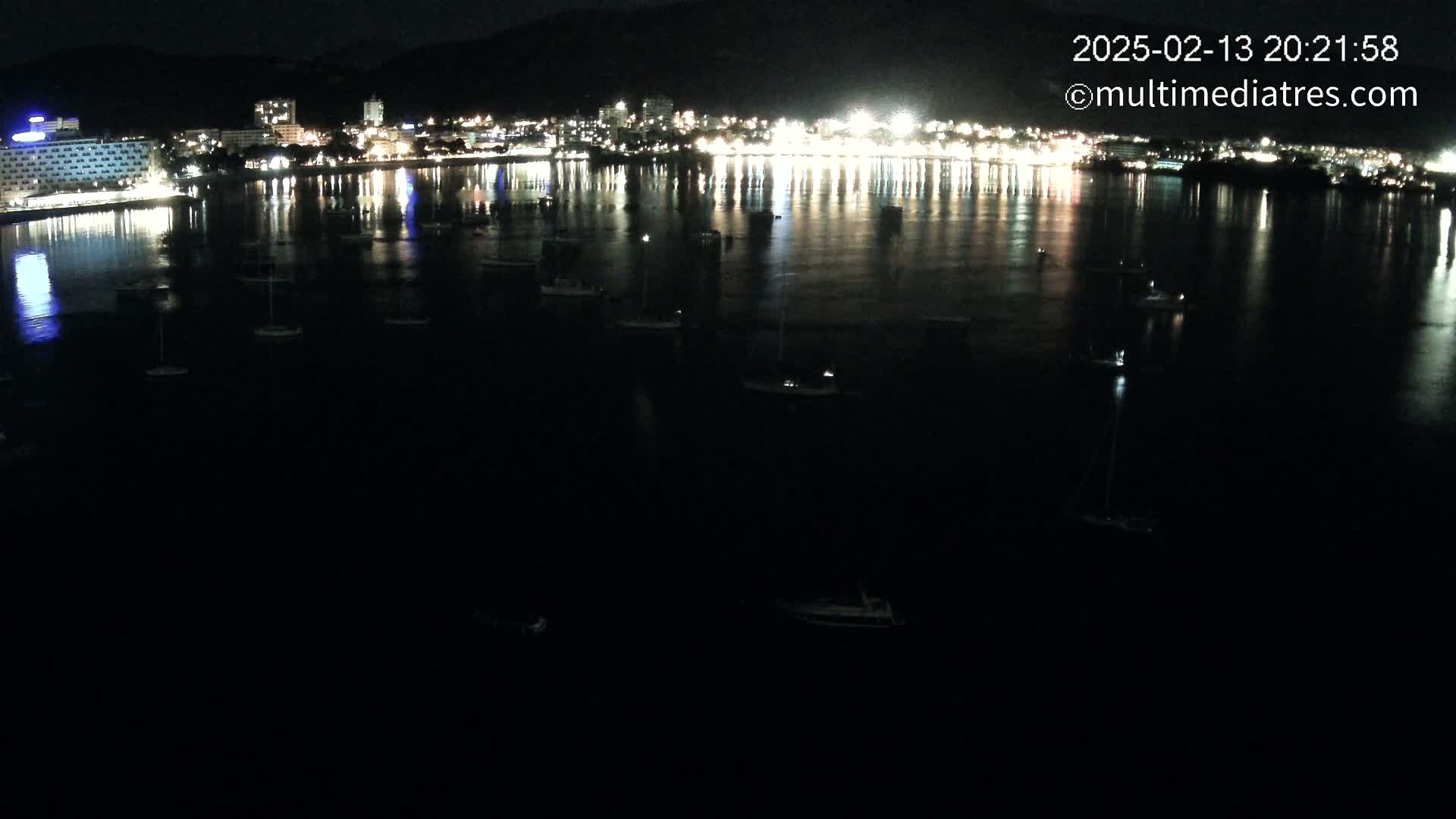 At night, a calm body of water reflects the lights of a city and numerous small boats are scattered across its surface.