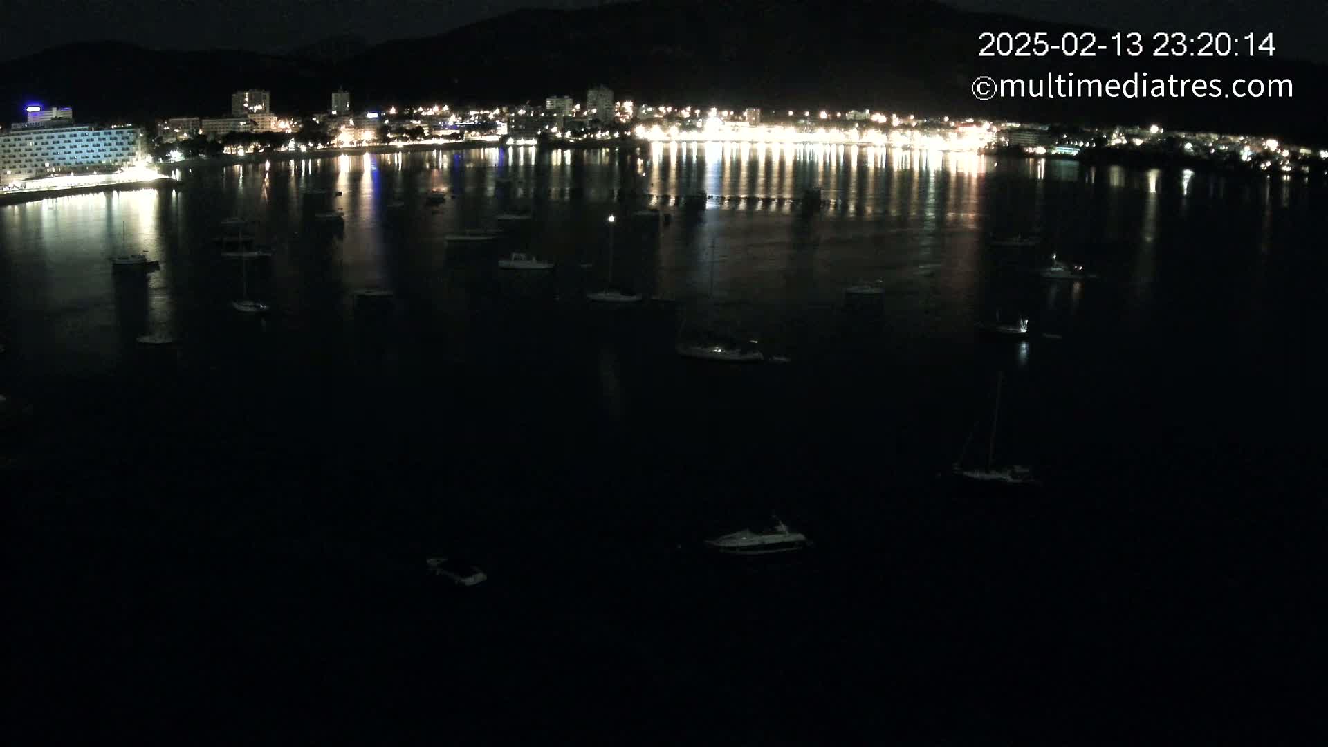 A nighttime aerial view shows a dark body of water with several small boats and a city's lights reflected on the water's surface.