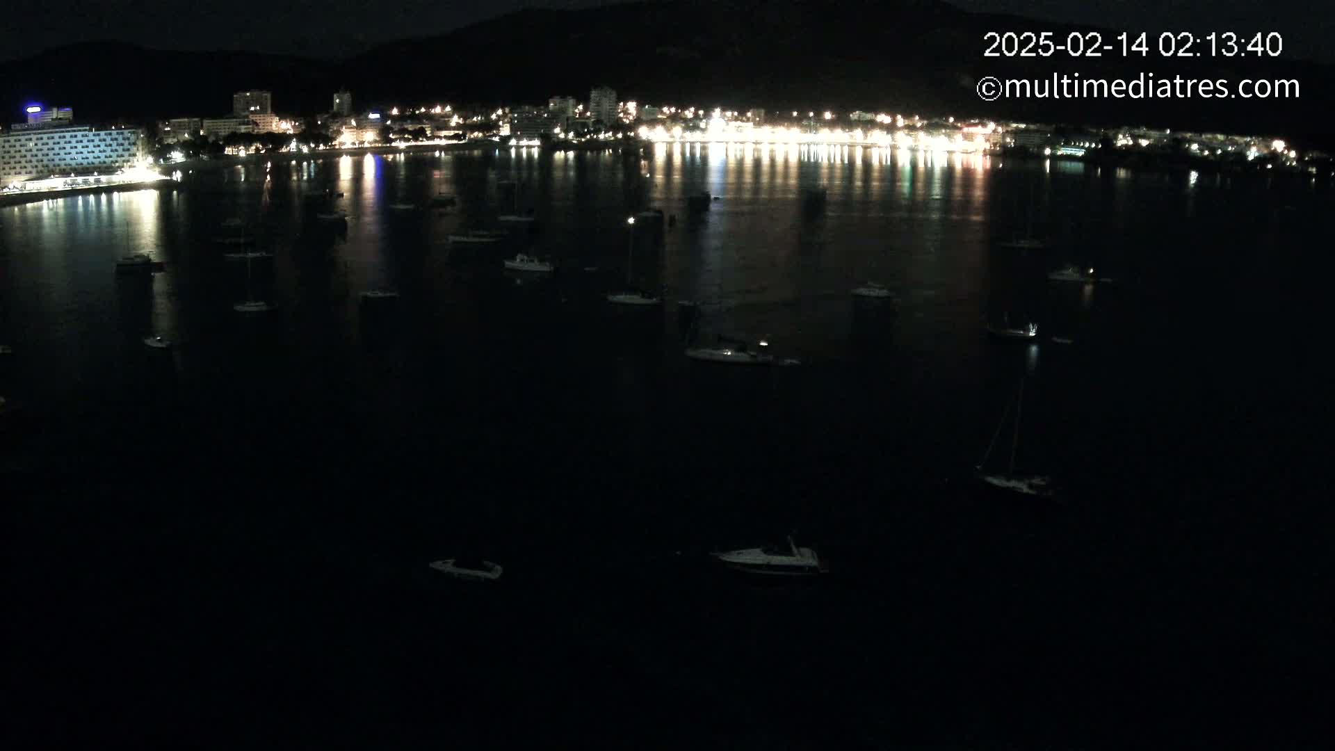 A nighttime view of a calm bay with numerous small boats scattered across its dark surface, reflecting the lights of a brightly lit town on the shore.