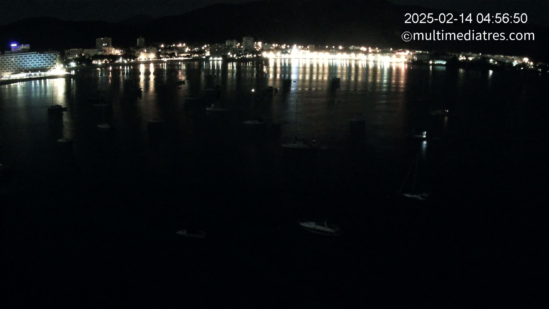 A nighttime, dark-water harbor scene shows a brightly lit town reflected in the water, with several boats scattered across the surface.