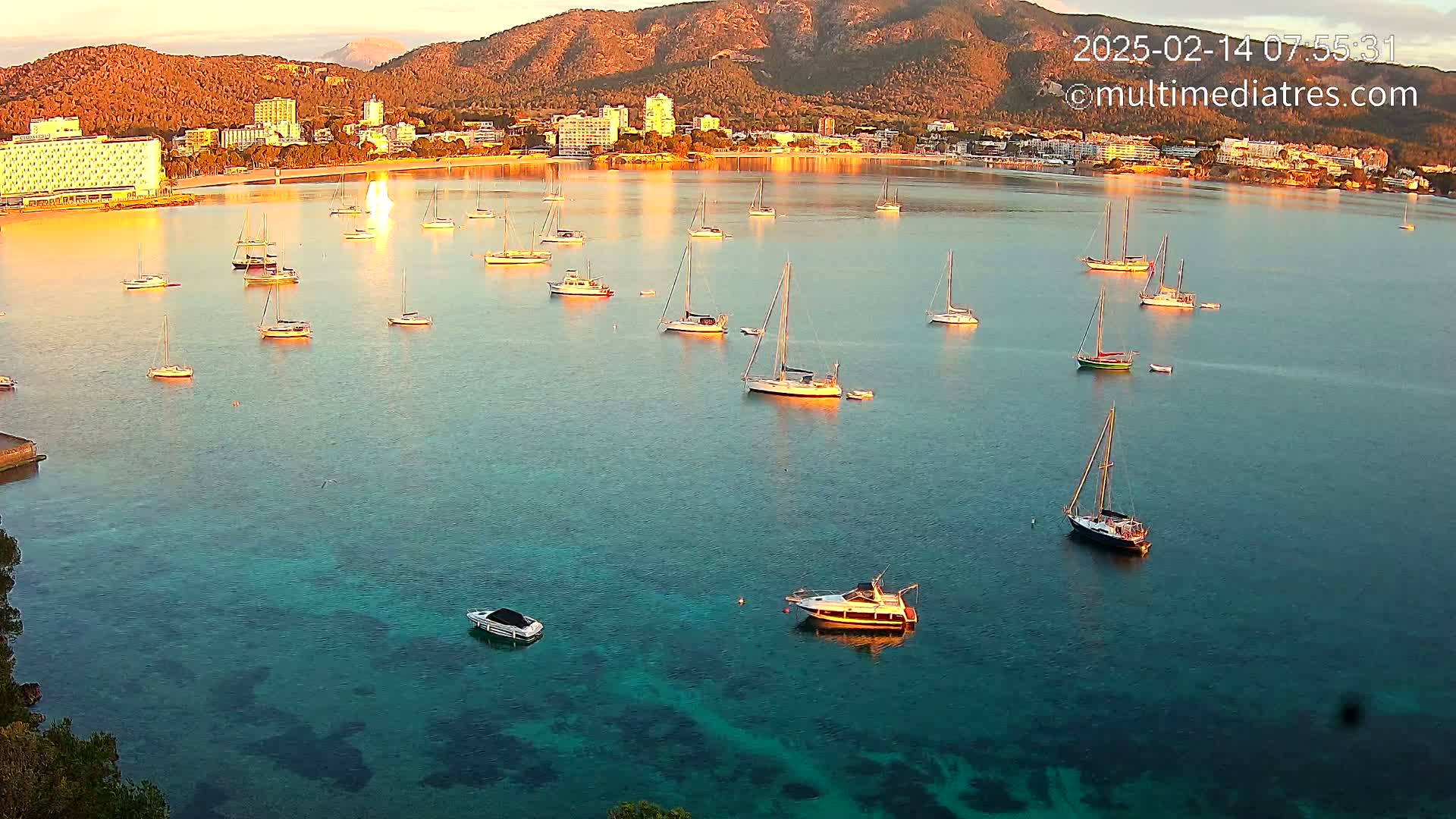 Numerous sailboats and motorboats are anchored in a calm, clear, teal-colored bay at sunrise, with a town and mountains visible in the background.