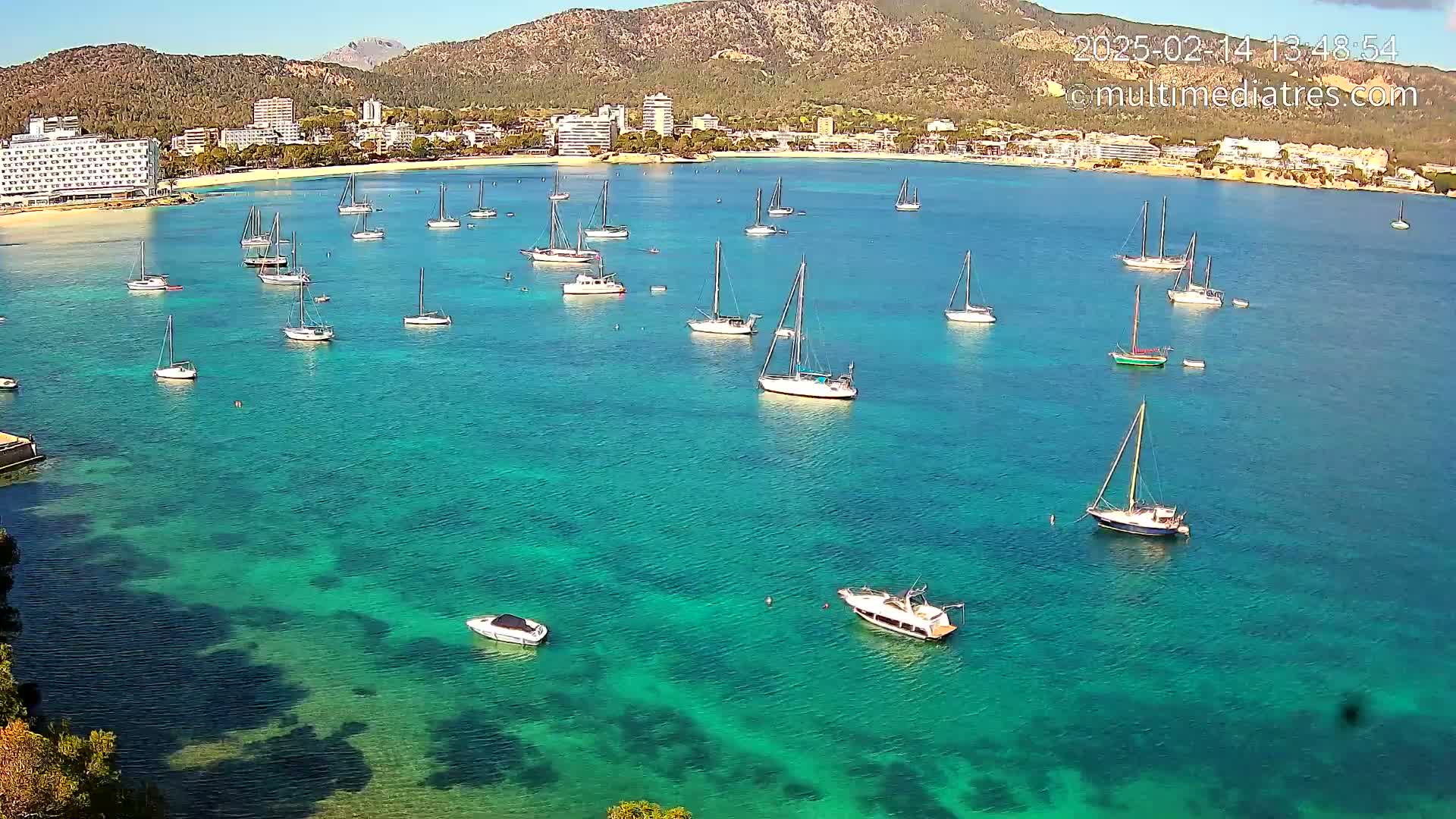 Numerous sailboats are anchored in a calm, clear turquoise bay near a coastline with hotels and mountains under a sunny sky.