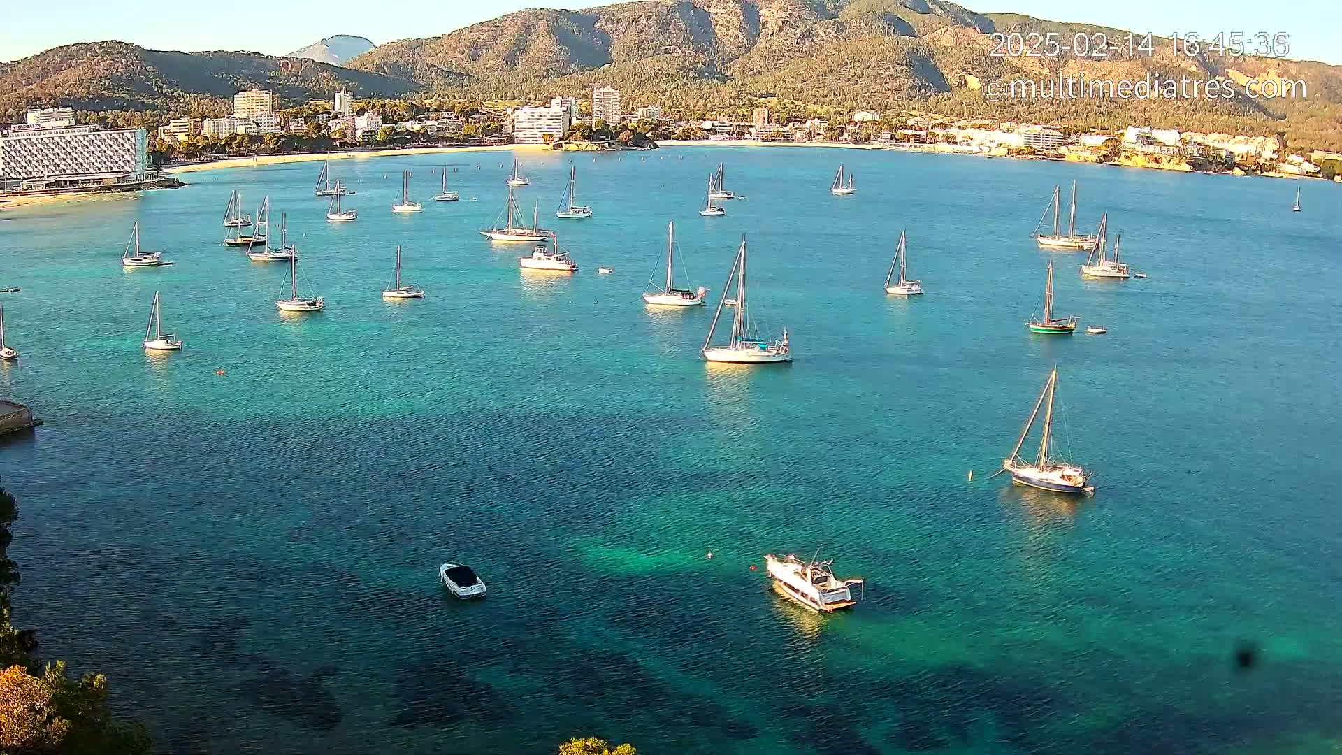 Numerous sailboats are anchored in a calm, teal bay near a coastline with buildings and mountains in the background.