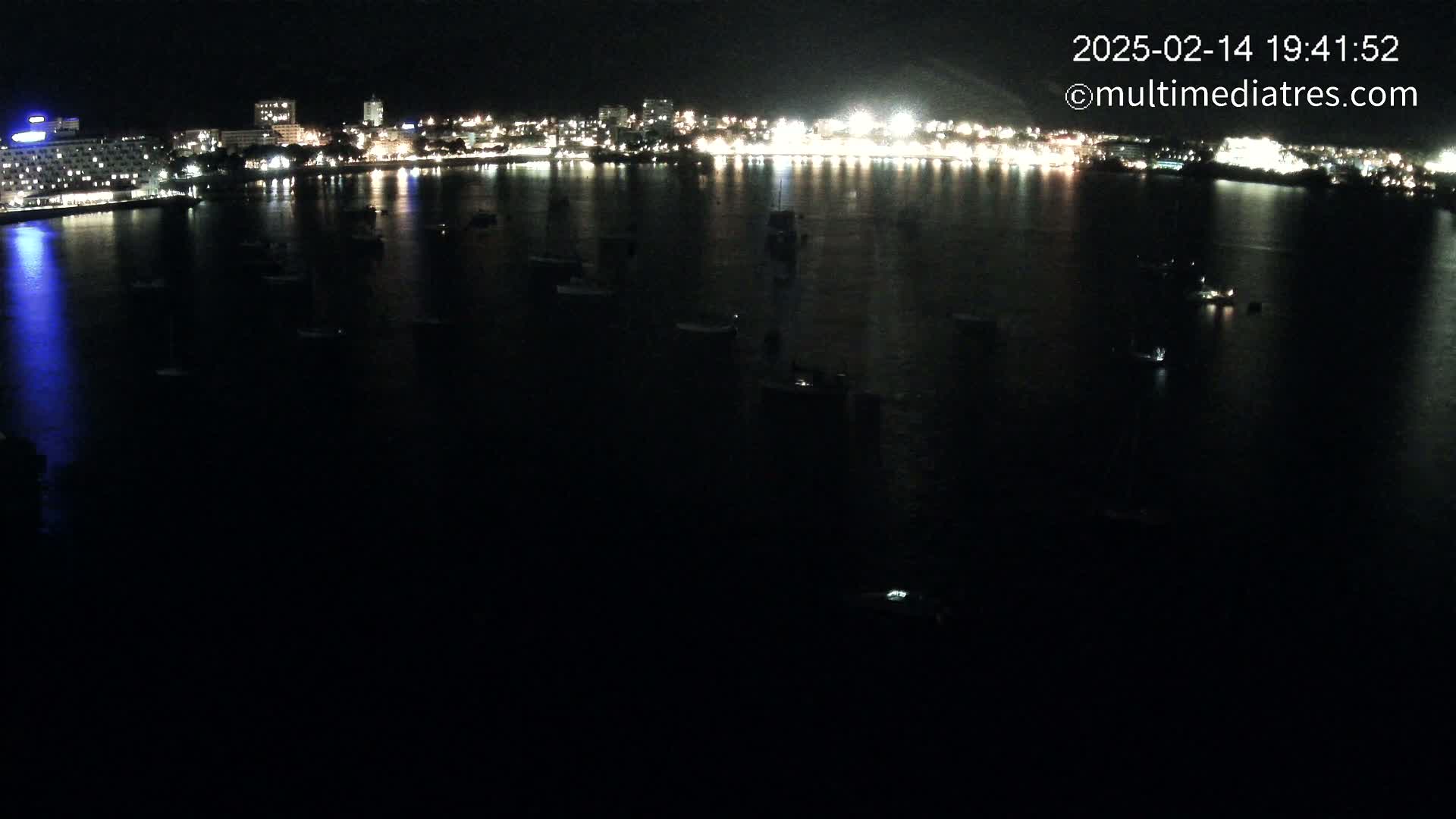 A nighttime view of a city's illuminated waterfront, with numerous boats scattered across a dark body of water.