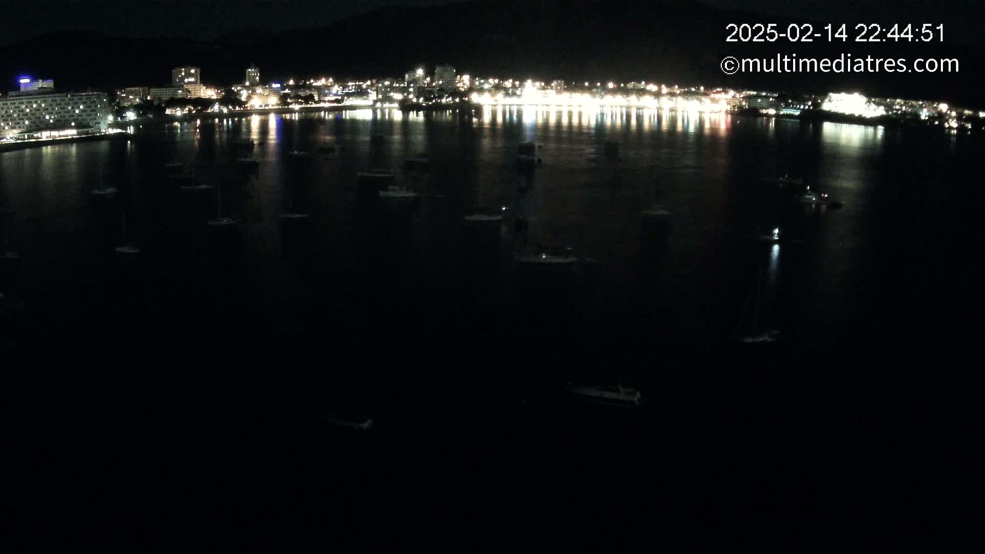 A nighttime, dark-water view of numerous small boats moored in a bay, with a brightly lit cityscape visible across the water under a clear sky.